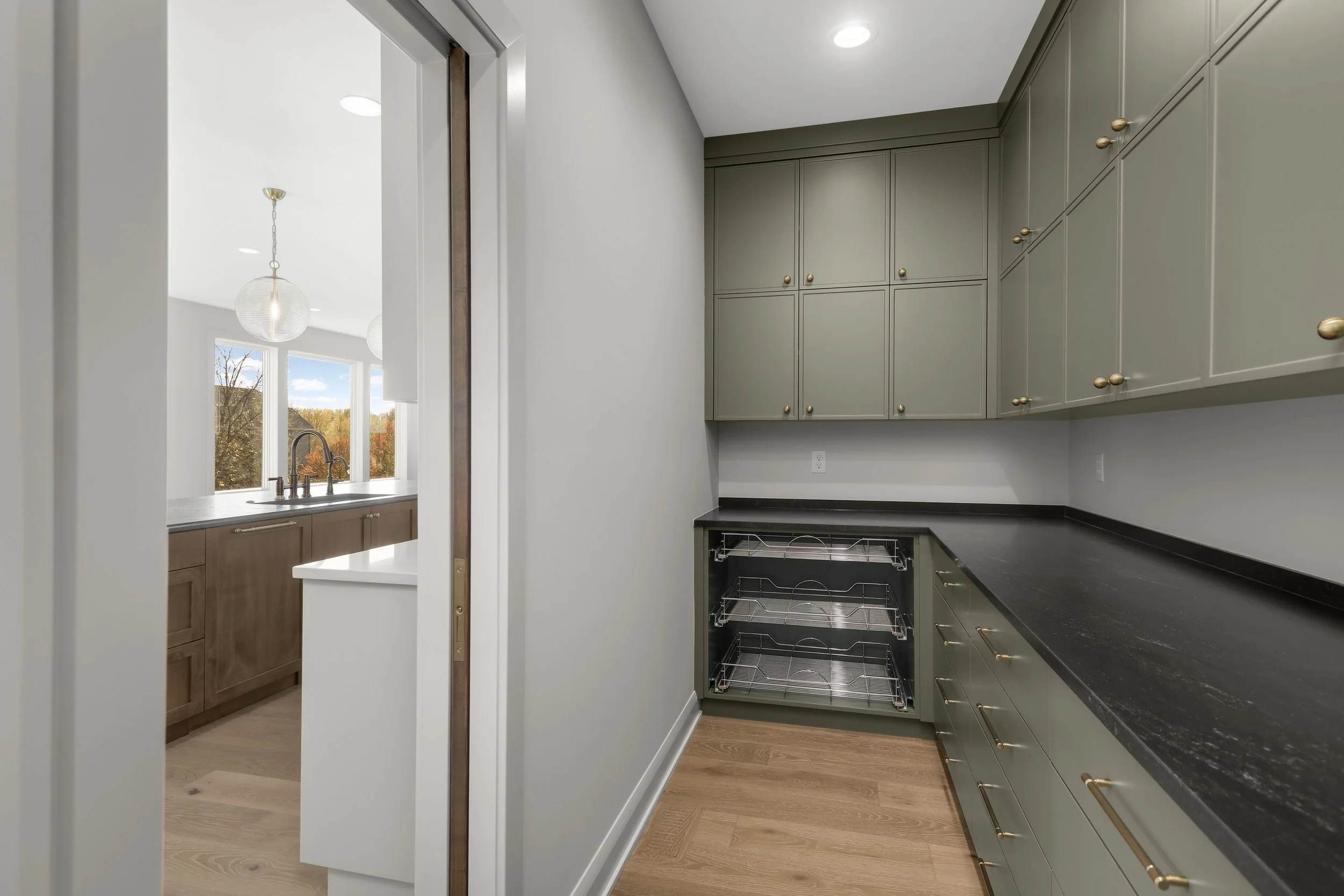 Modern galley kitchen with gray cabinets, black countertops, brass hardware, wood flooring, and a pull-out pantry beside a bright sink area.