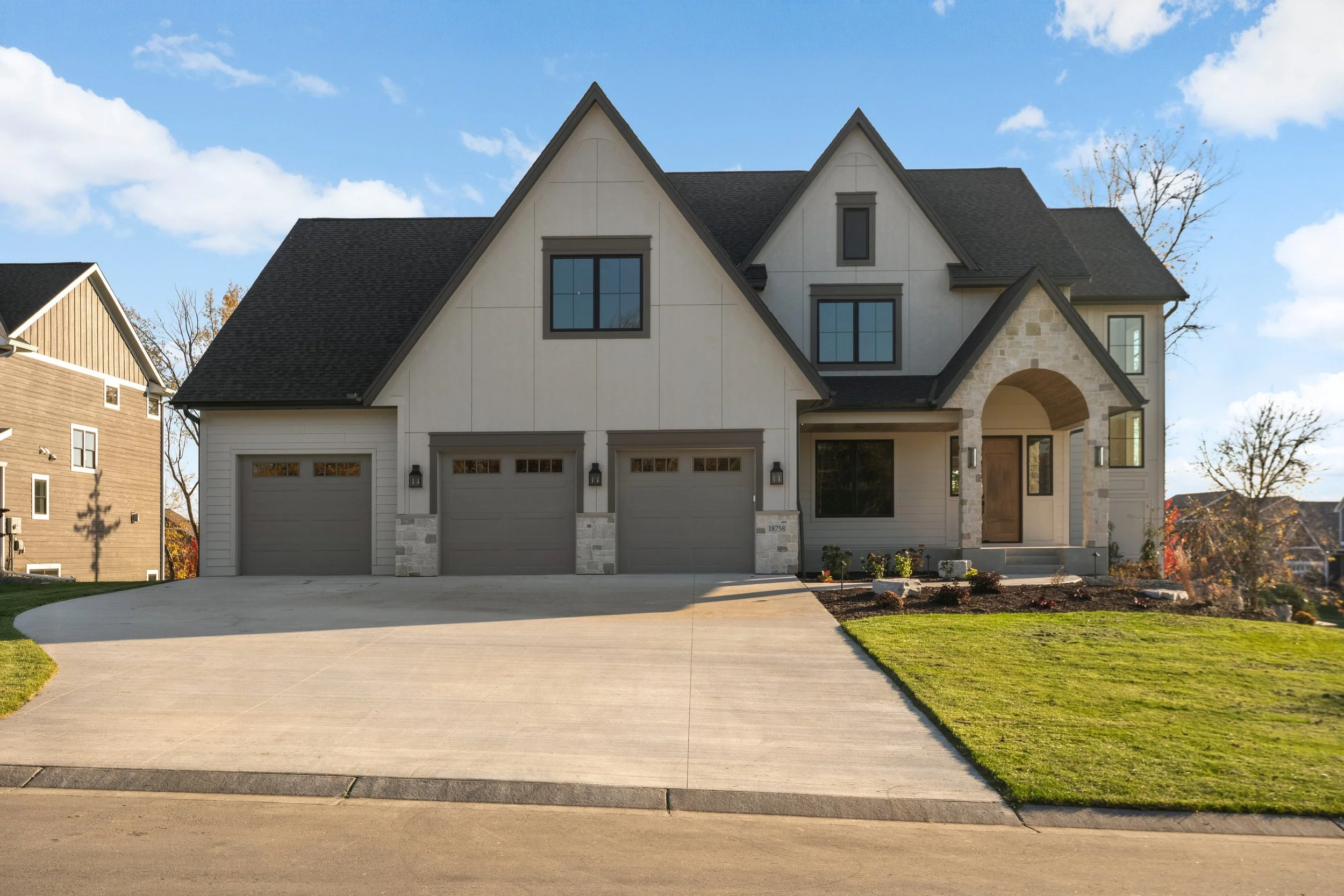 Large modern suburban house with three-car garage, stone entryway, steep gabled roof, driveway, and landscaped lawn
