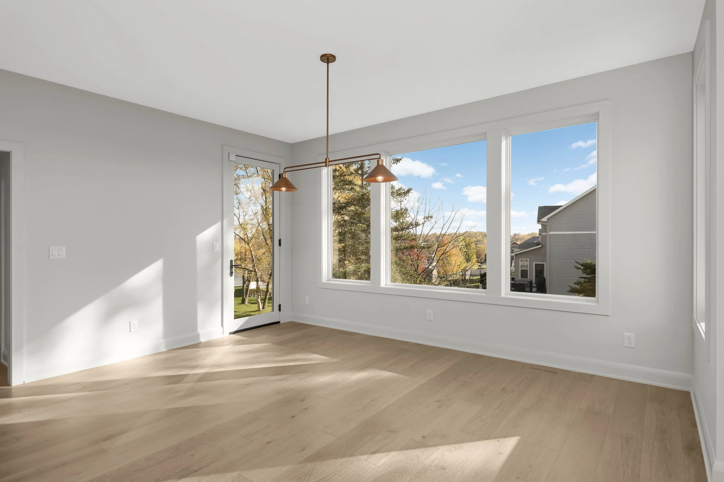 Bright empty room with light wood floors, large windows, glass door, and modern bronze pendant lights