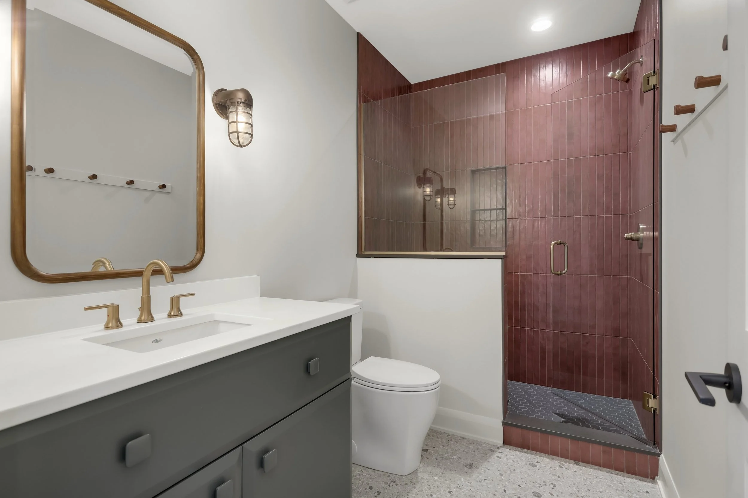Modern bathroom with gray vanity, white countertop, wood-framed mirror, toilet, and burgundy tiled walk-in shower.