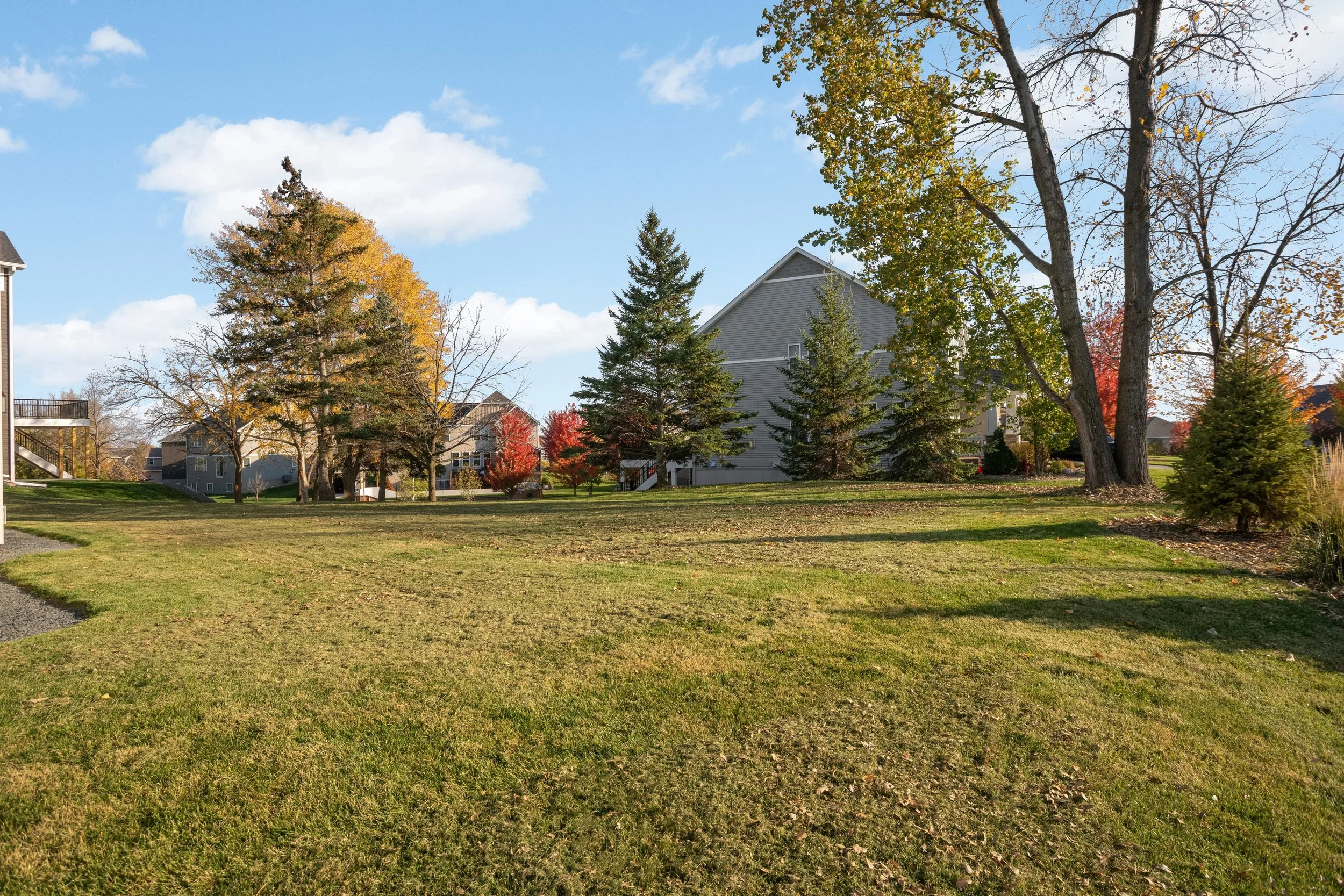 Suburban neighborhood with gray homes, green lawn, evergreen trees, and colorful autumn foliage under a blue sky