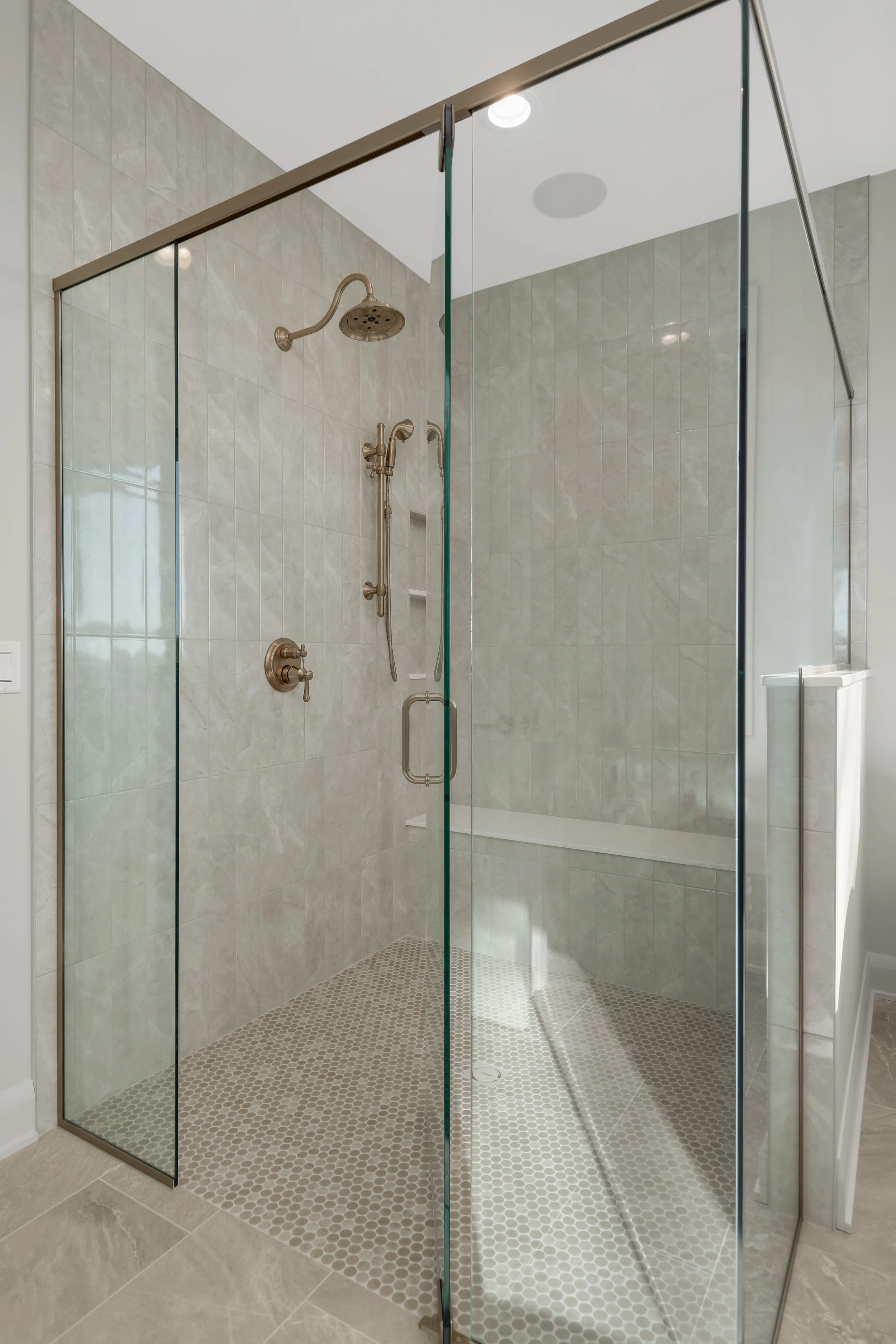 Modern walk-in bathroom shower with frameless glass doors, marble-look tile walls, handheld and rainfall showerheads, built-in bench, and penny tile floor.
