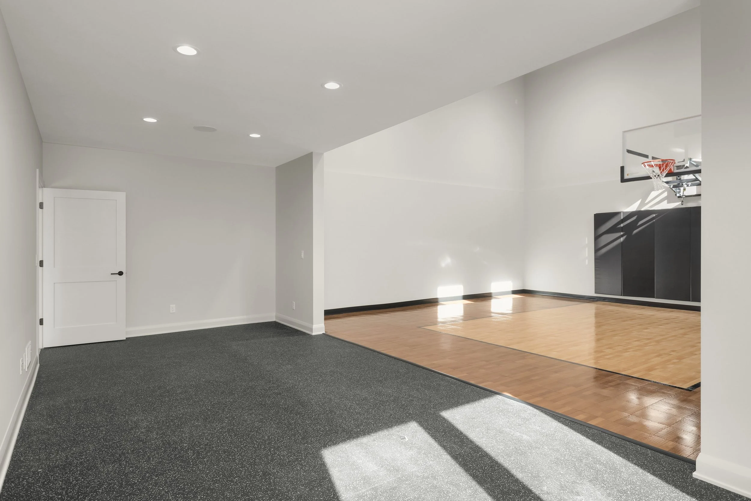 Indoor basketball court with hardwood flooring, basketball hoop, black wall padding, and adjacent carpeted area