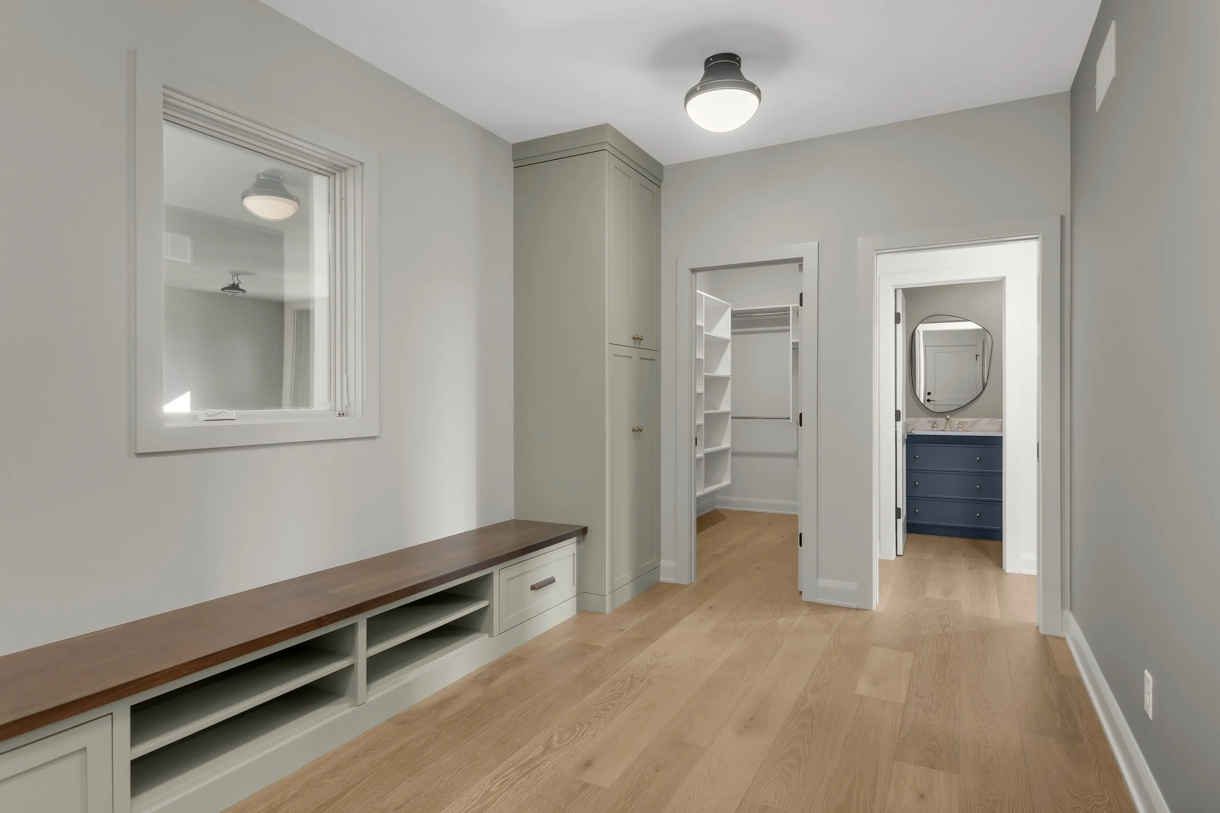 Bright modern hallway with built-in white cabinetry, wood bench, hardwood floors, walk-in closet, and bathroom beyond