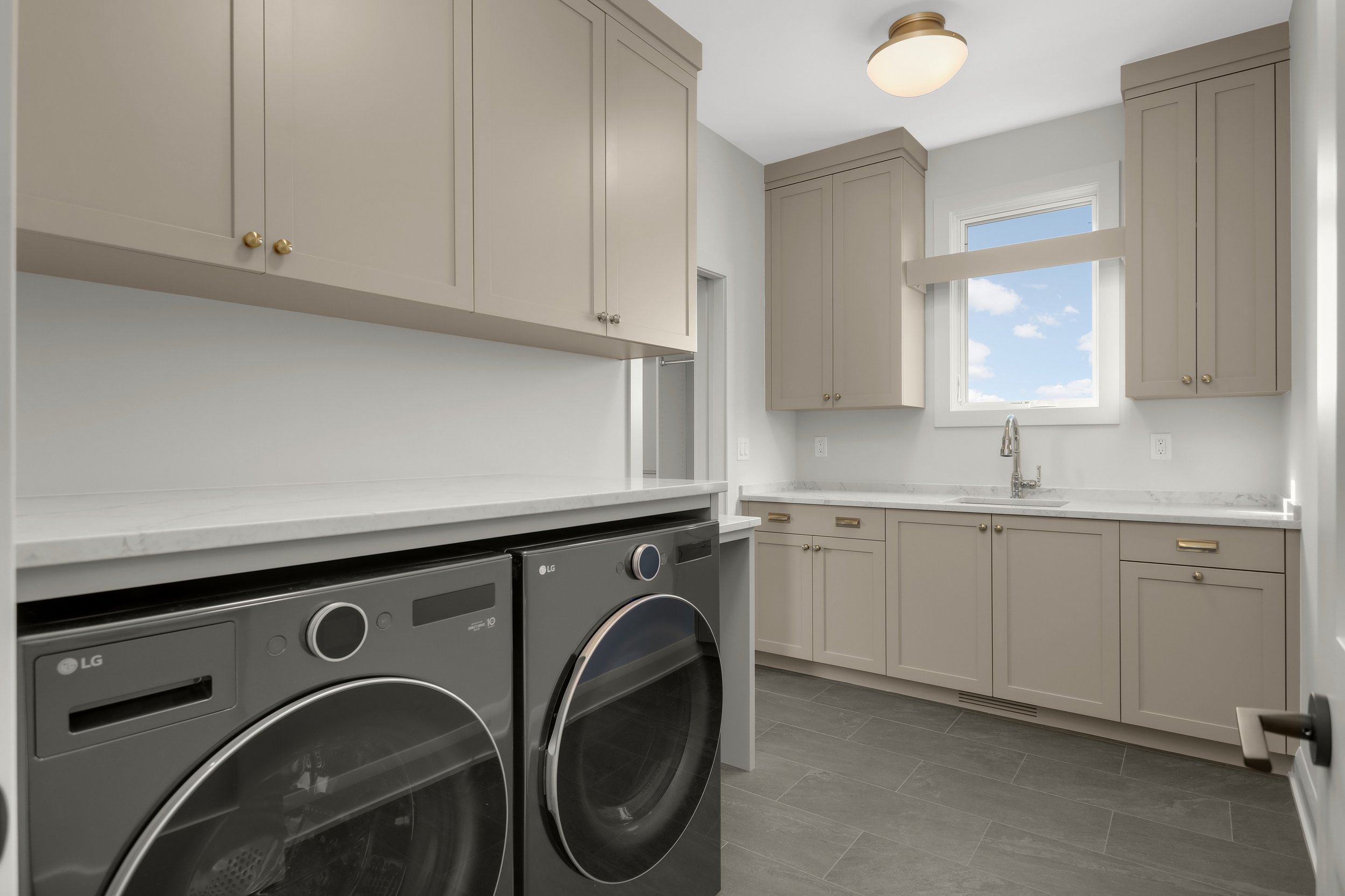 Modern laundry room with gray cabinets, white countertops, LG front-loading washer and dryer, utility sink, and window