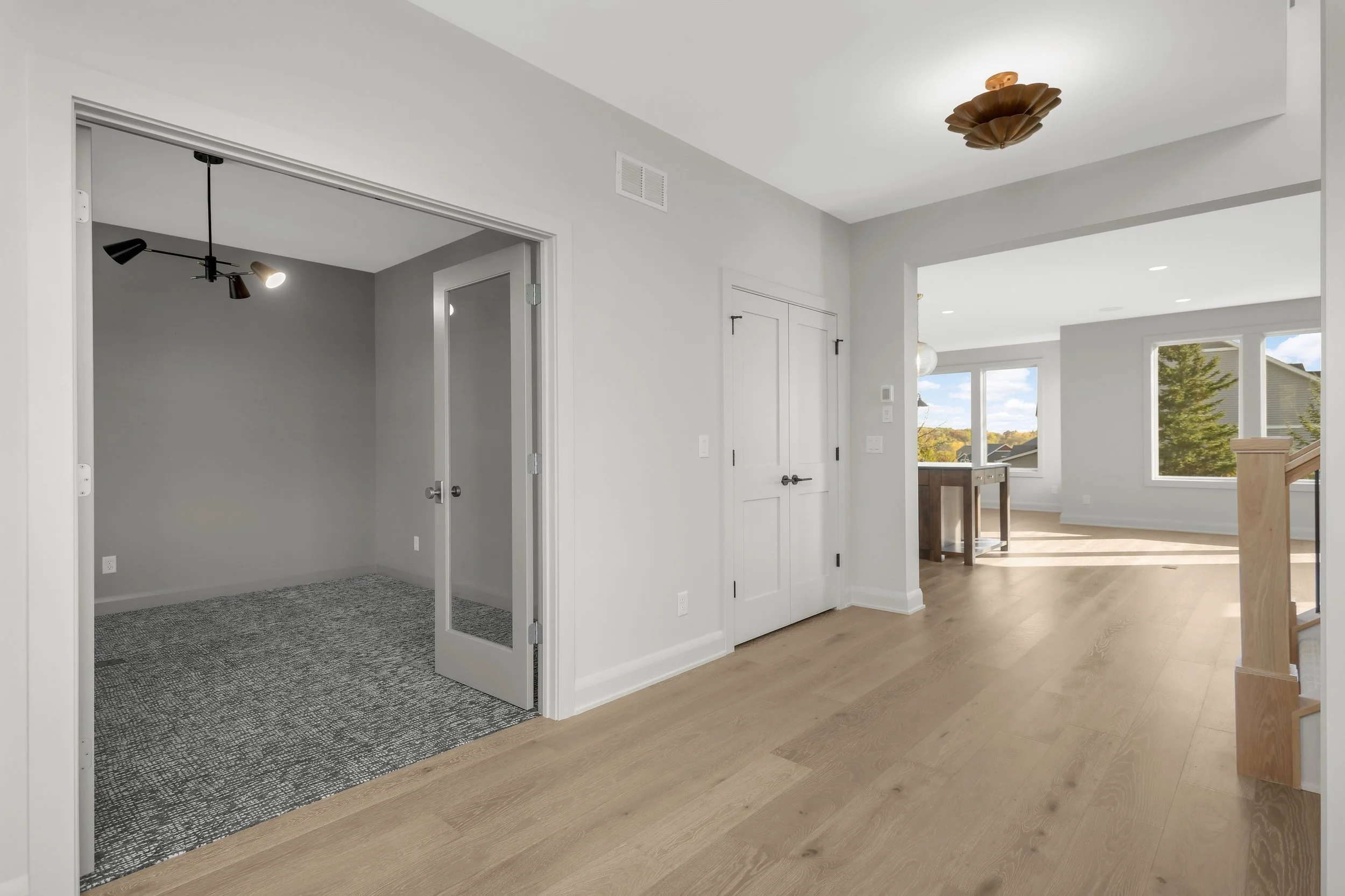 Bright modern home hallway with hardwood floors, double doors, carpeted room, decorative ceiling light, staircase, and sunlit living area