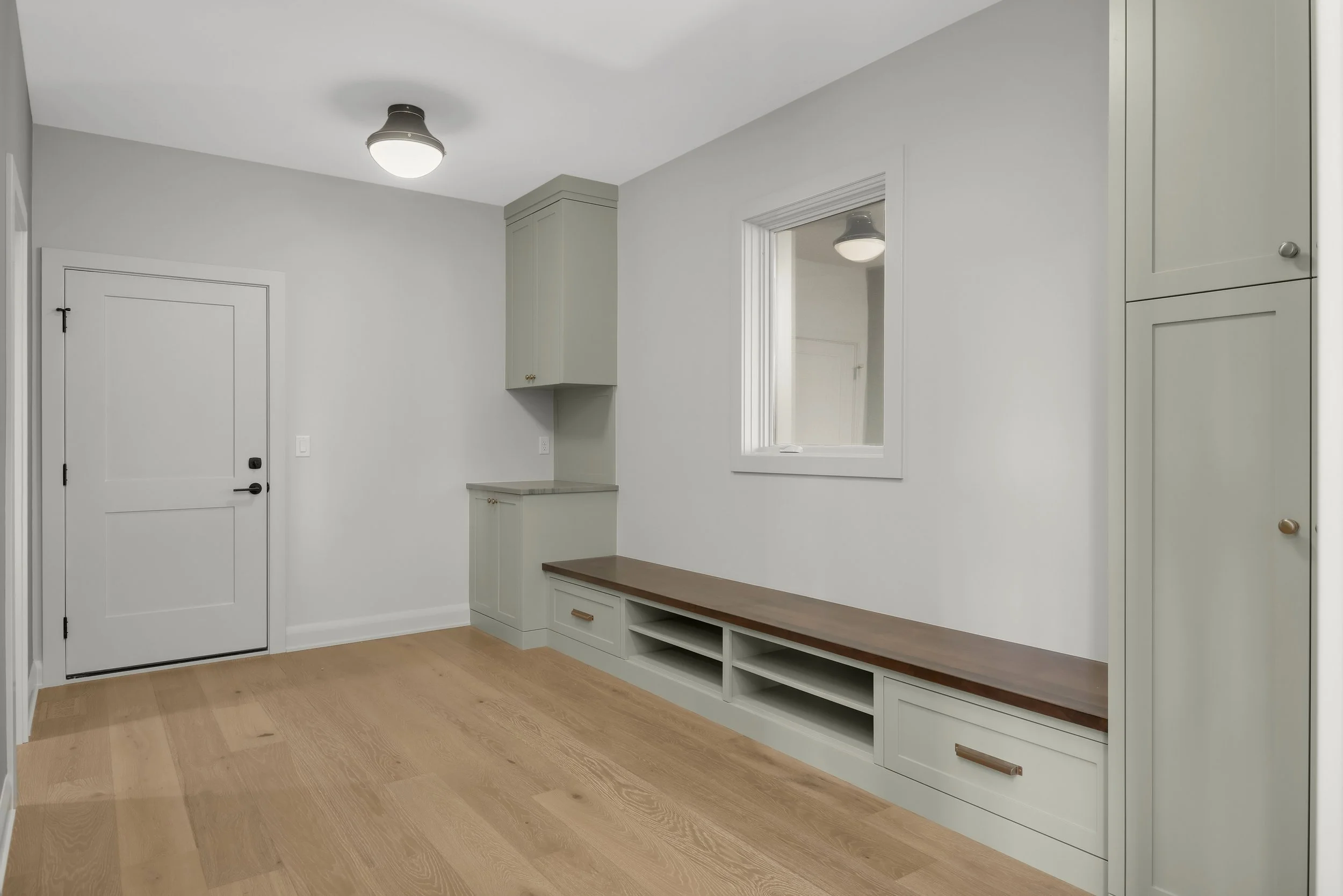 Bright entryway with built-in gray cabinetry, wood bench, storage drawers, mirror, white door, and light wood flooring