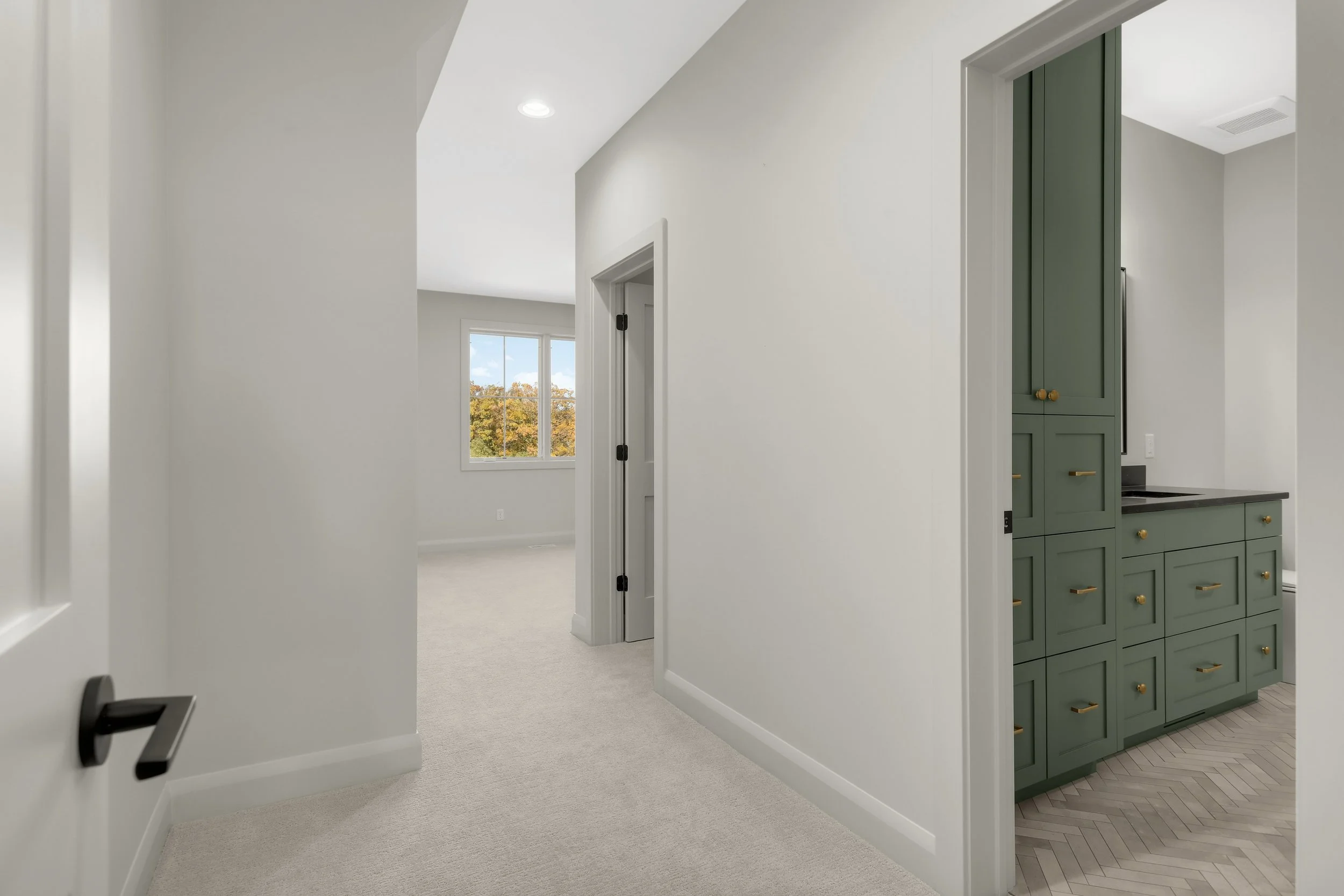 Bright home hallway with carpeted floors, a bedroom window, and a bathroom featuring green cabinetry, brass hardware, and herringbone tile flooring.