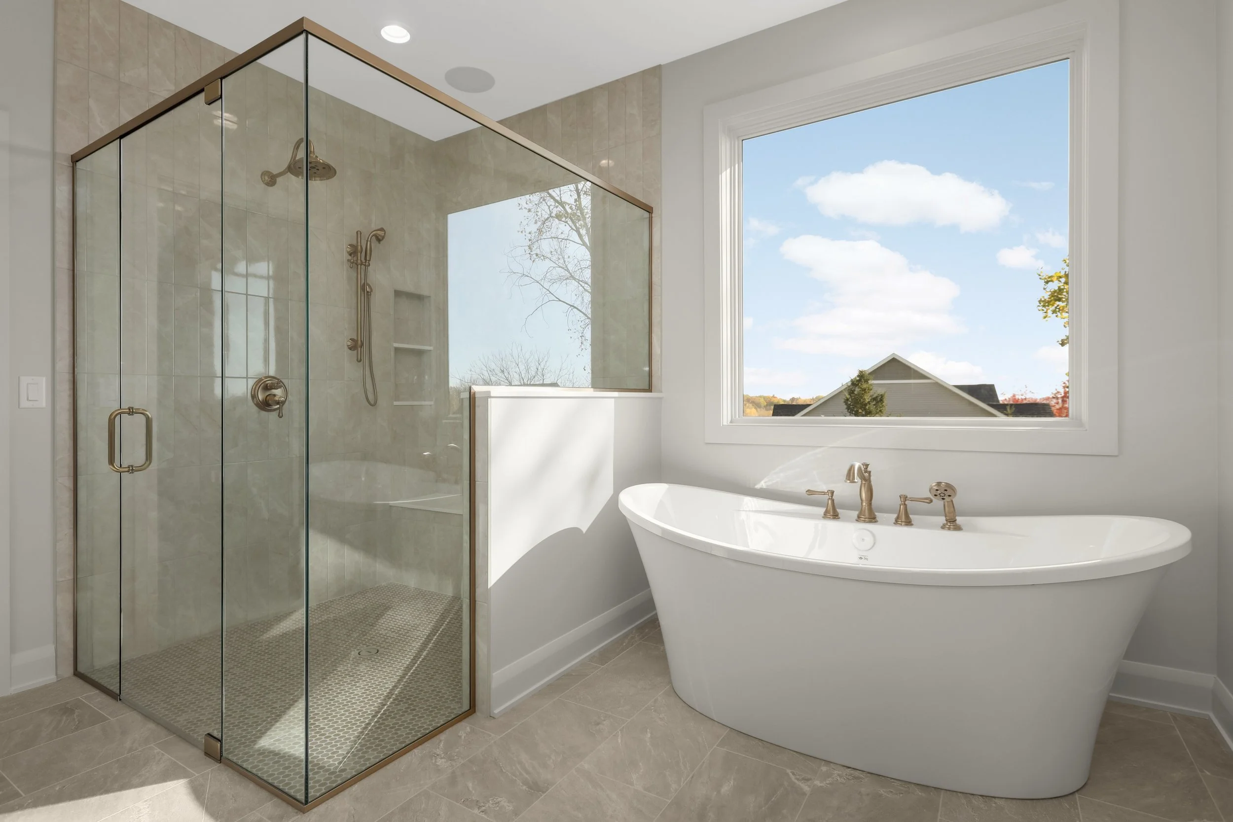 Bright modern bathroom with a glass-enclosed walk-in shower, freestanding soaking tub, beige tile, and large window