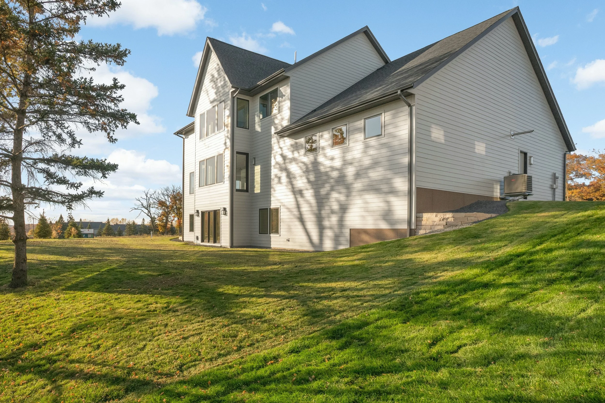 Modern white-sided house with multiple gabled roofs, large windows, and a lush sloping lawn under a blue sky