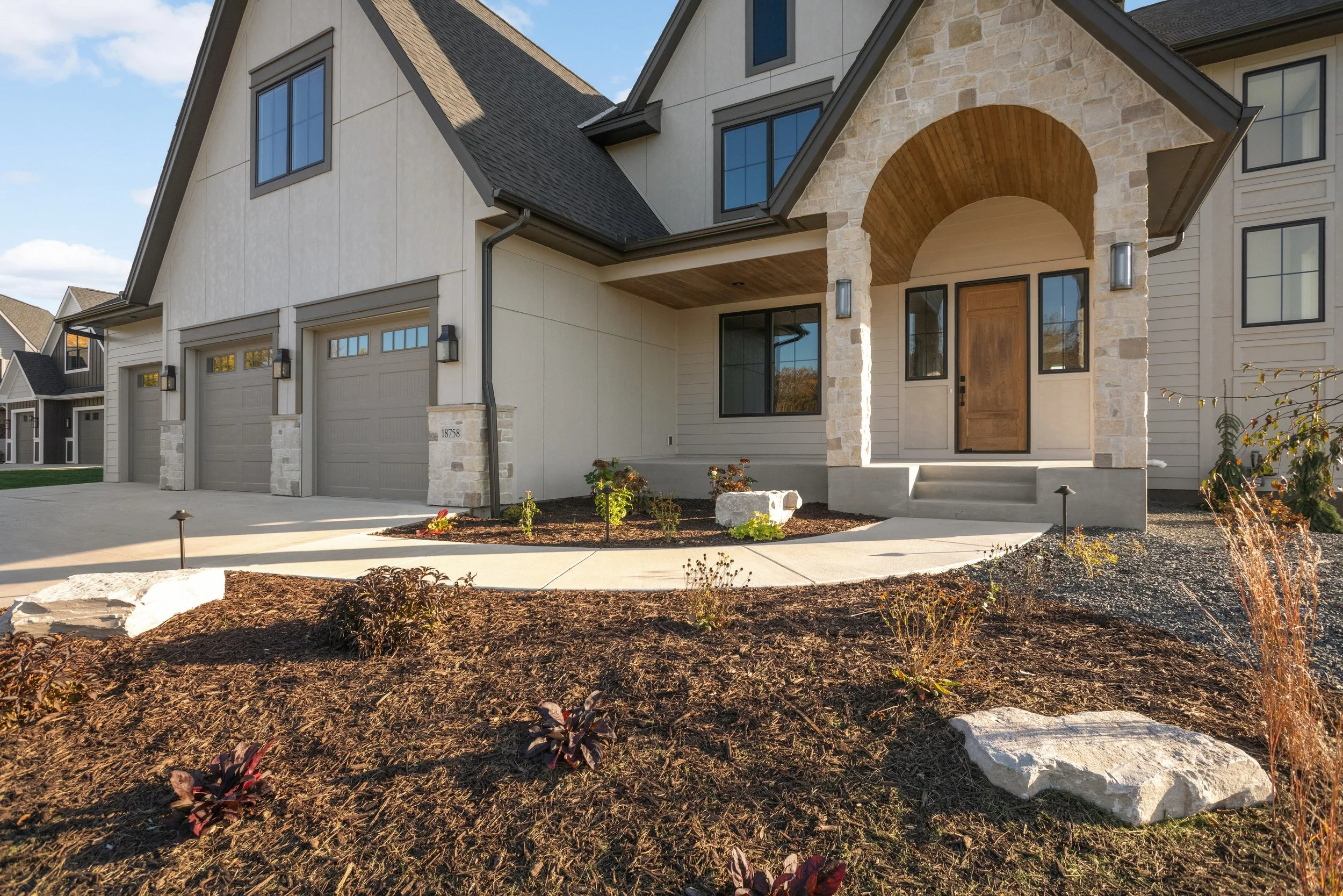 Modern suburban home exterior with stone-arched entryway, wooden front door, double garage, landscaped yard, and concrete walkway