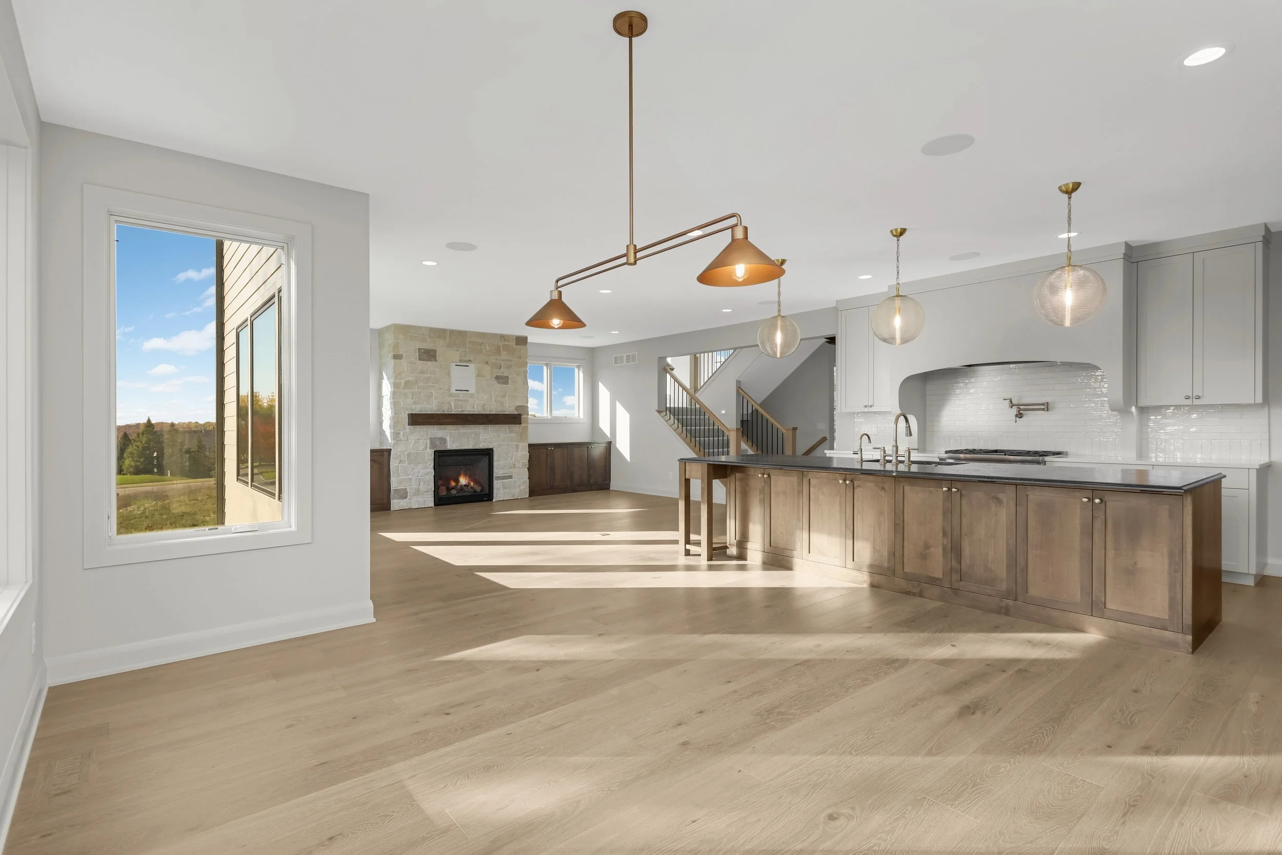 Bright modern open-concept kitchen and living room with a large island, pendant lighting, stone fireplace, hardwood floors, and staircase