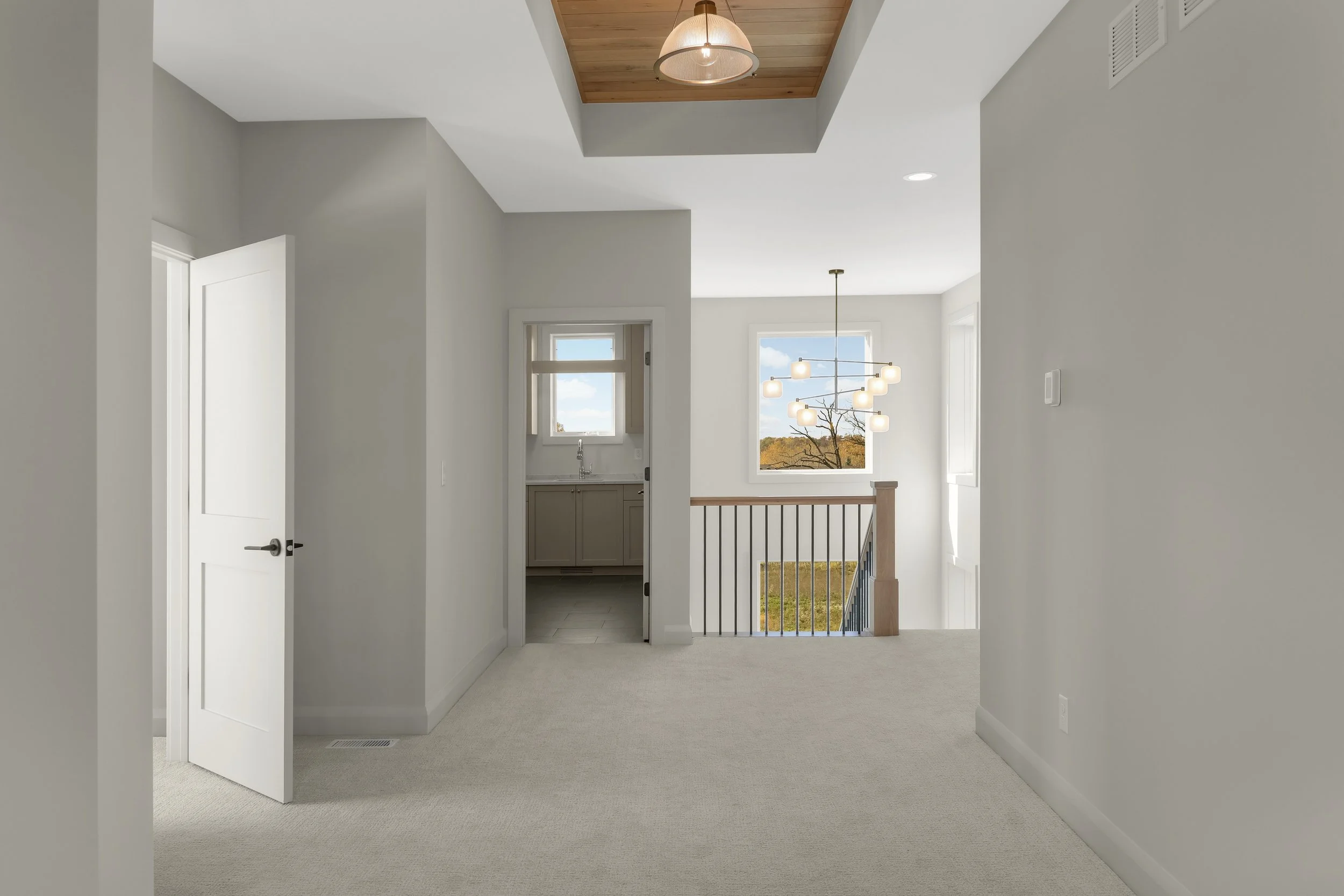 Bright modern upstairs hallway with carpeted floors, open white door, laundry room, wood-paneled ceiling, pendant lights, windows, and stair railing.