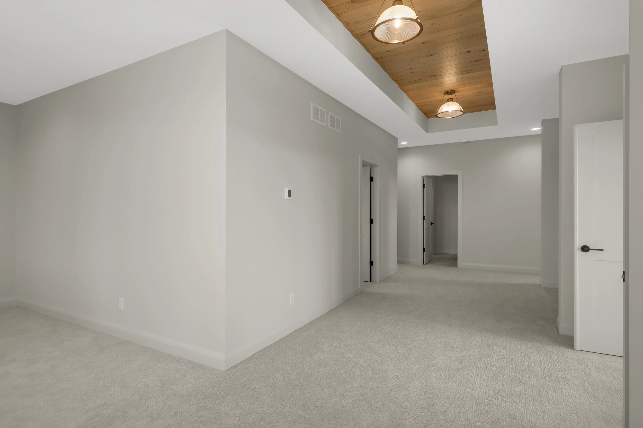 Spacious modern carpeted hallway with white walls, multiple doorways, and wood-paneled ceiling with pendant lights