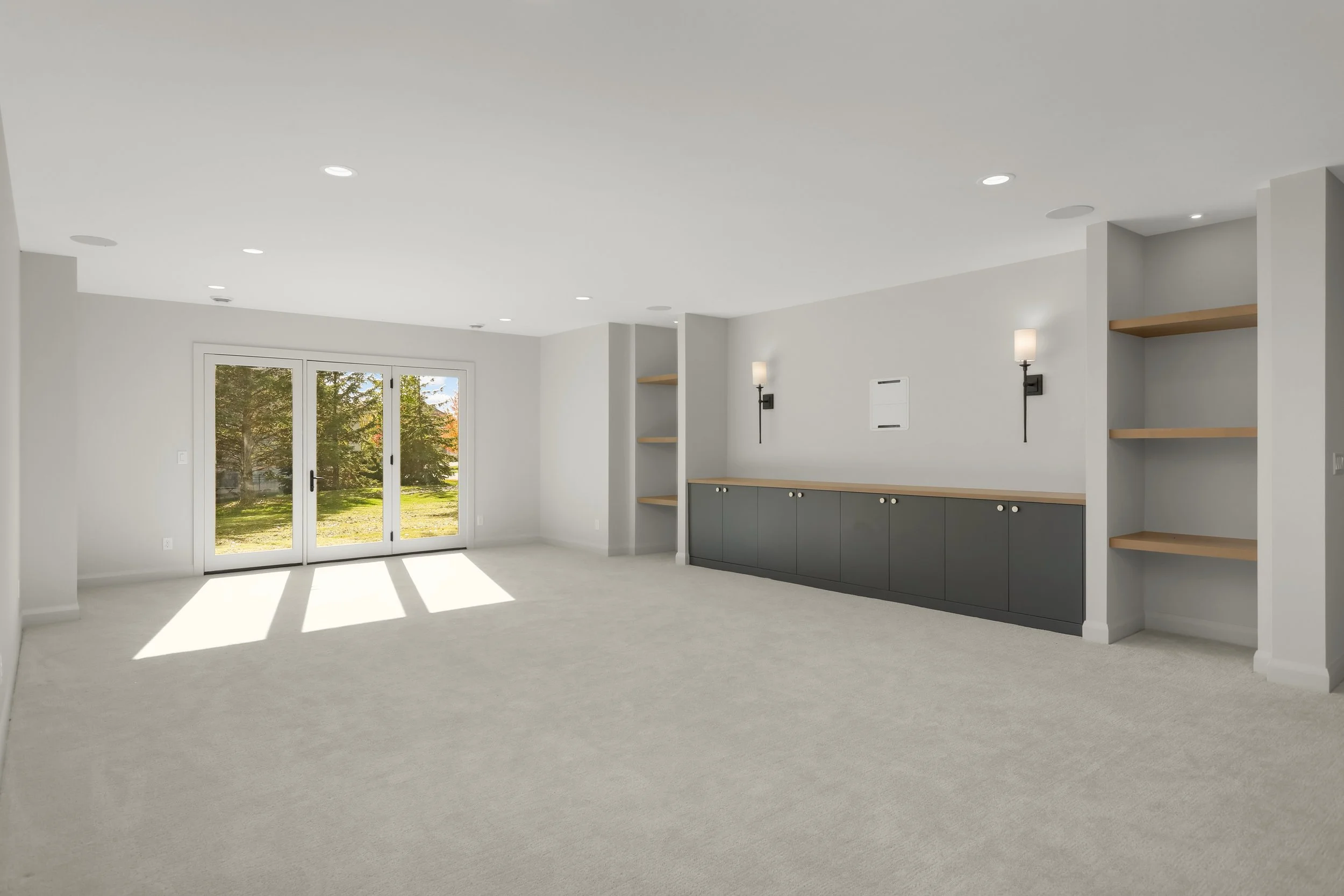 Bright modern living room with built-in dark cabinets, wood shelving, wall sconces, recessed lighting, and glass double doors opening to a landscaped yard