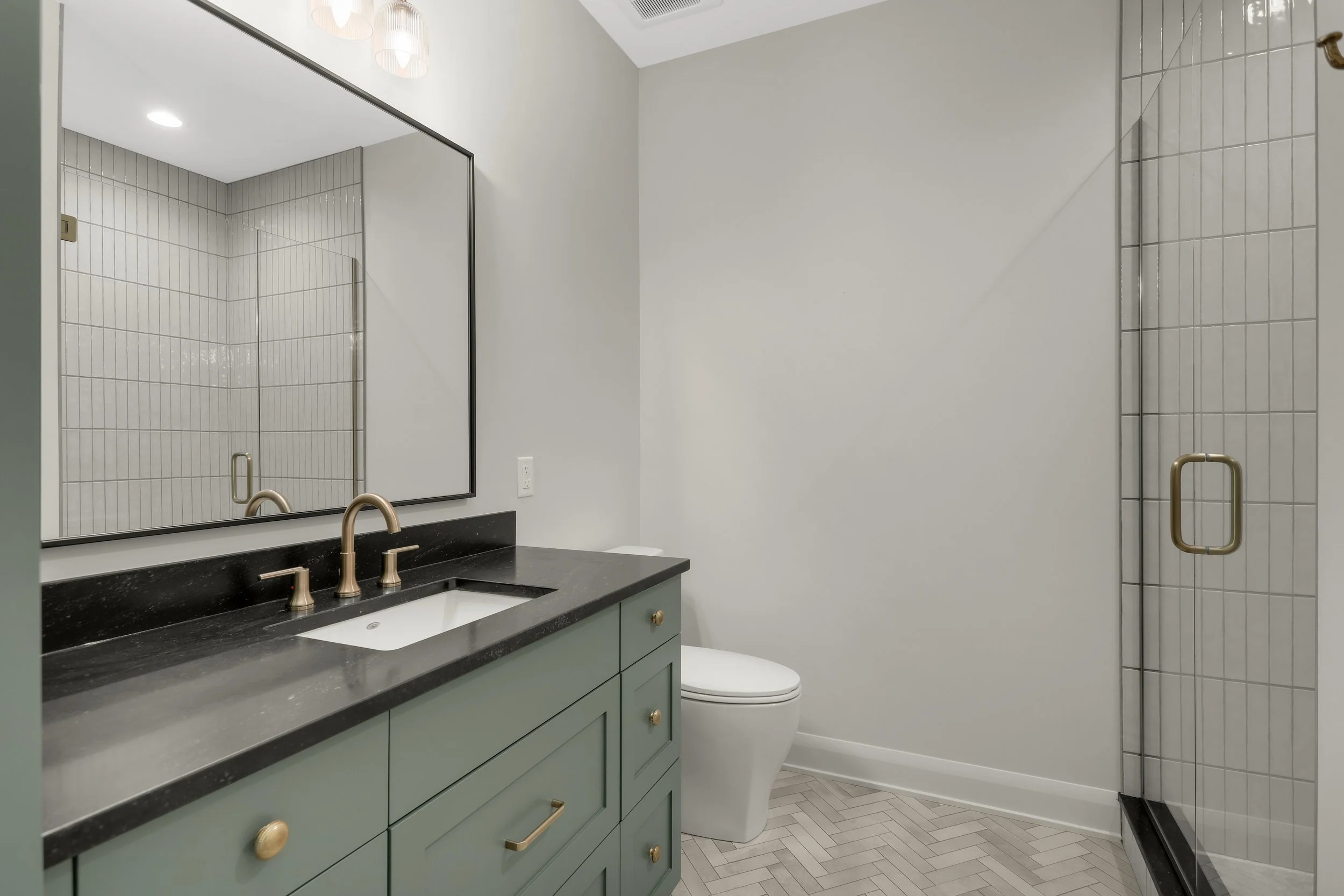 Modern bathroom with green vanity, black countertop, brass fixtures, large mirror, toilet, and white tiled shower.