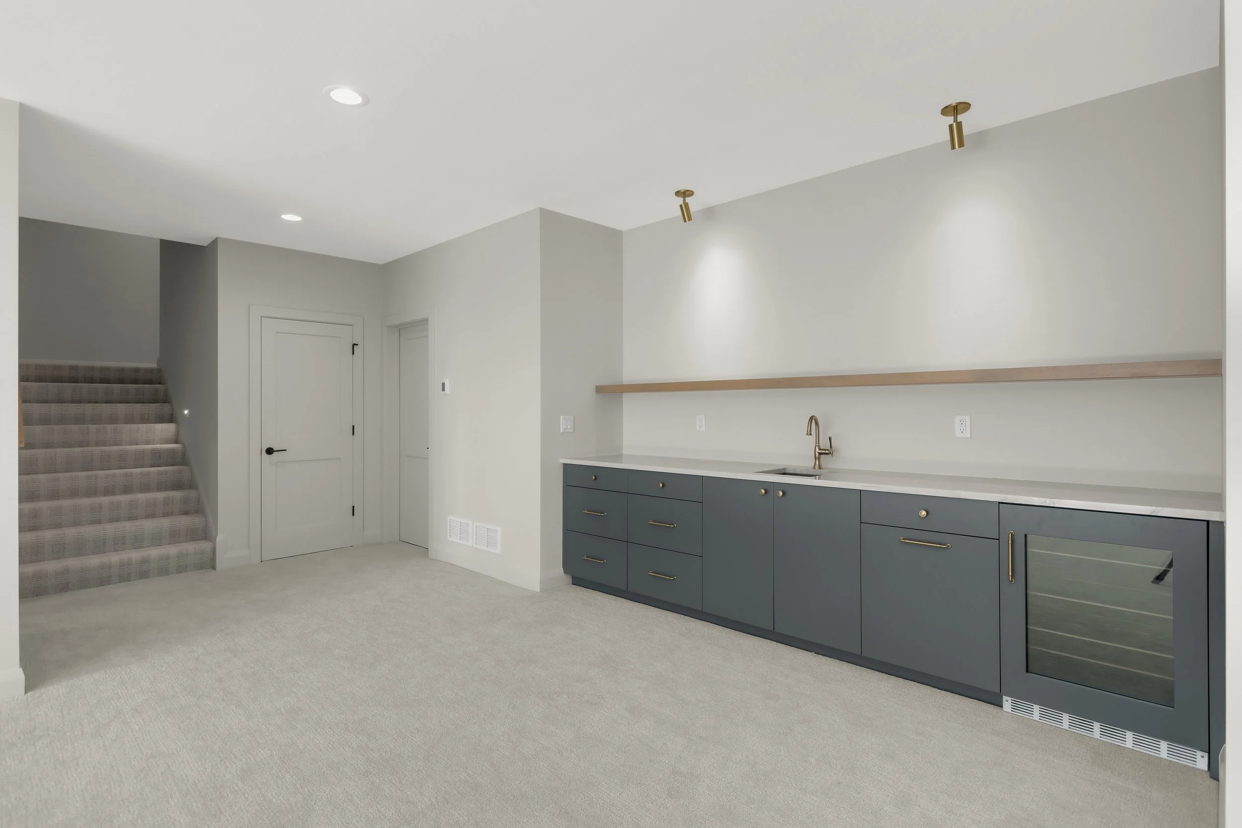Modern basement kitchenette with dark blue cabinets, white countertop, sink, beverage refrigerator, and carpeted staircase