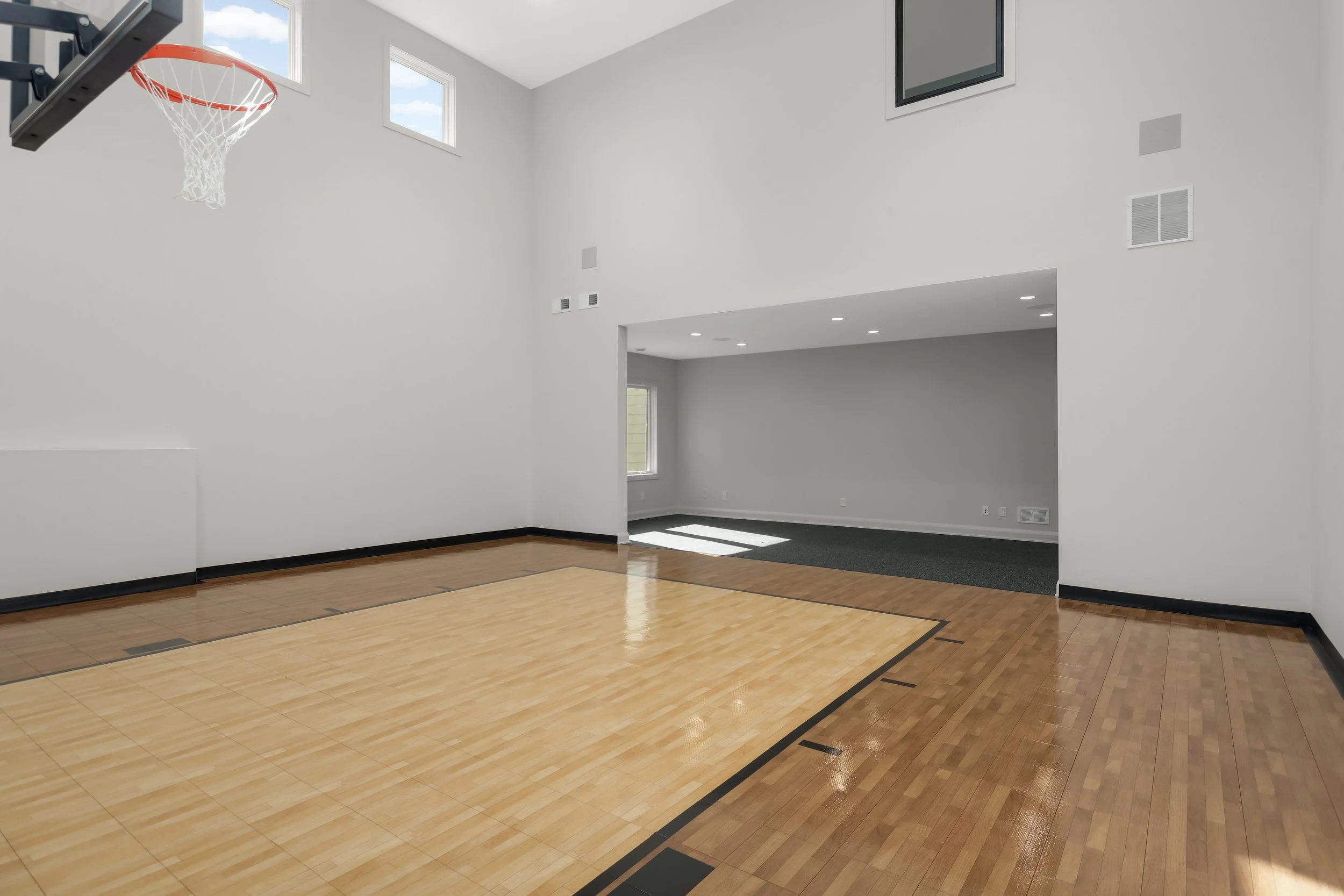 Indoor basketball court with polished hardwood flooring, hoop, high windows, and adjacent gym space