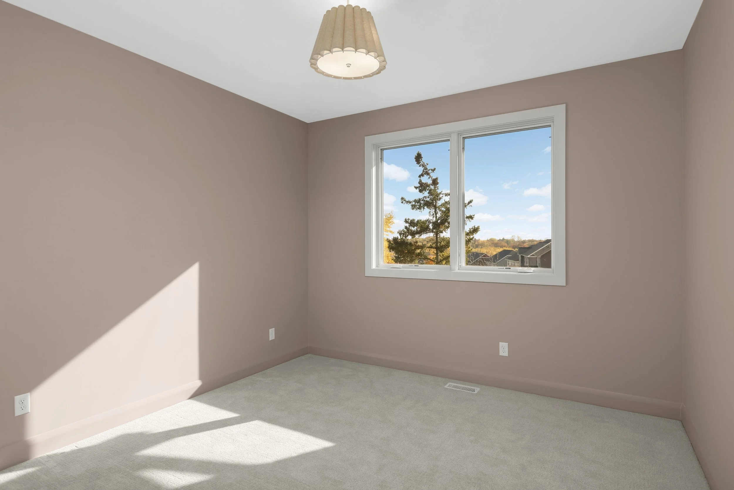 Empty carpeted bedroom with mauve walls, ceiling light fixture, and large window overlooking trees and rooftops