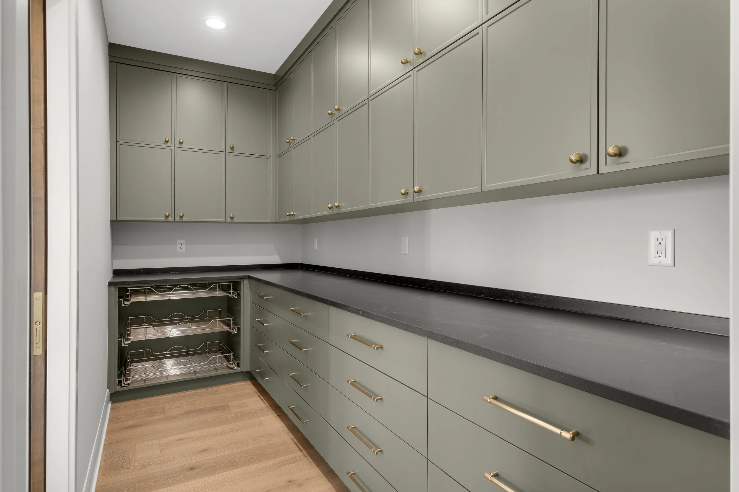 Walk-in pantry with gray cabinets, brass hardware, black countertops, and pull-out storage shelves