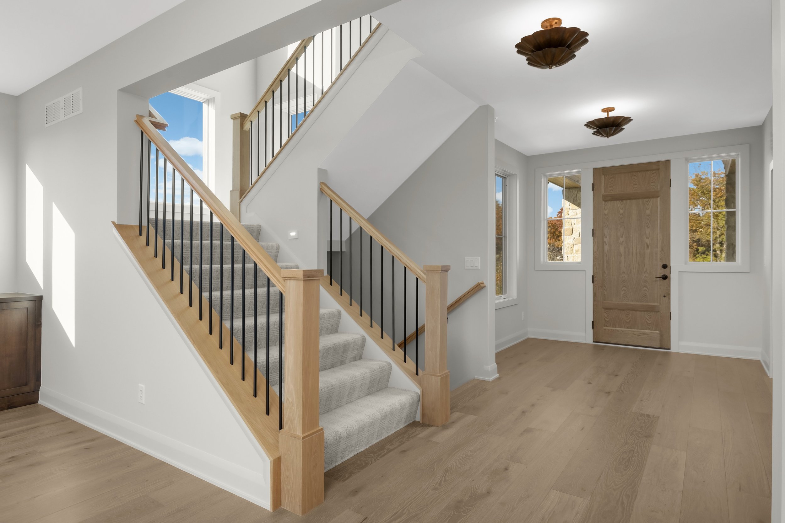 Bright modern home foyer with carpeted staircase, light wood railings, black metal balusters, hardwood floors, and a wooden front door