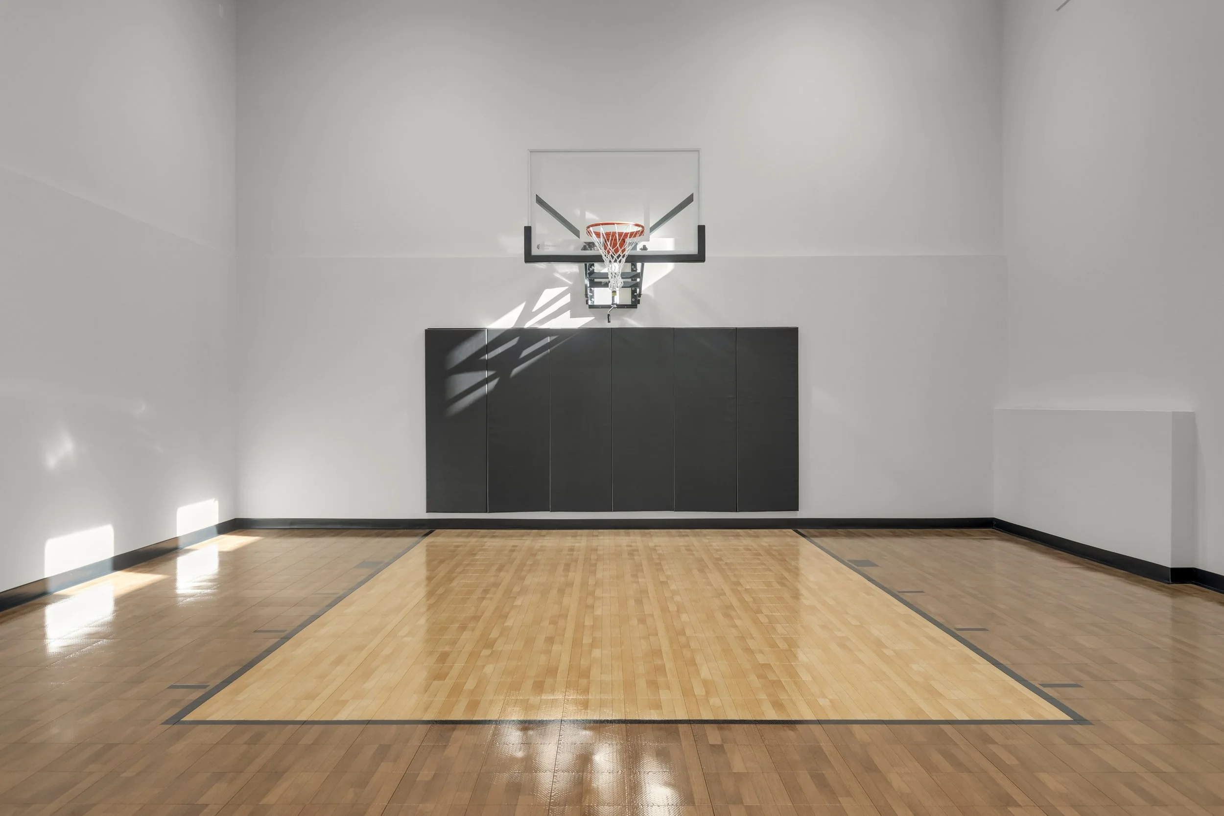 Empty indoor basketball court with a hoop, backboard, wall padding, and polished wood floor