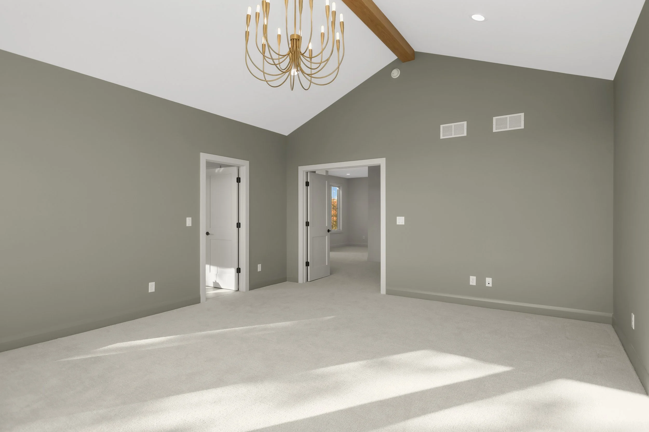 Empty gray-carpeted room with white trim, doors, recessed lighting, and a gold multi-arm chandelier
