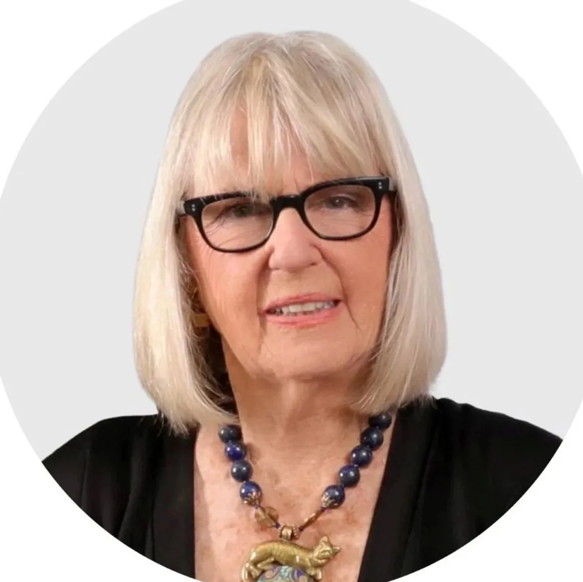A woman, Harriet Cohen, with blonde hair, black glasses, and a black top, wearing a blue and gold necklace with a fish pendant.