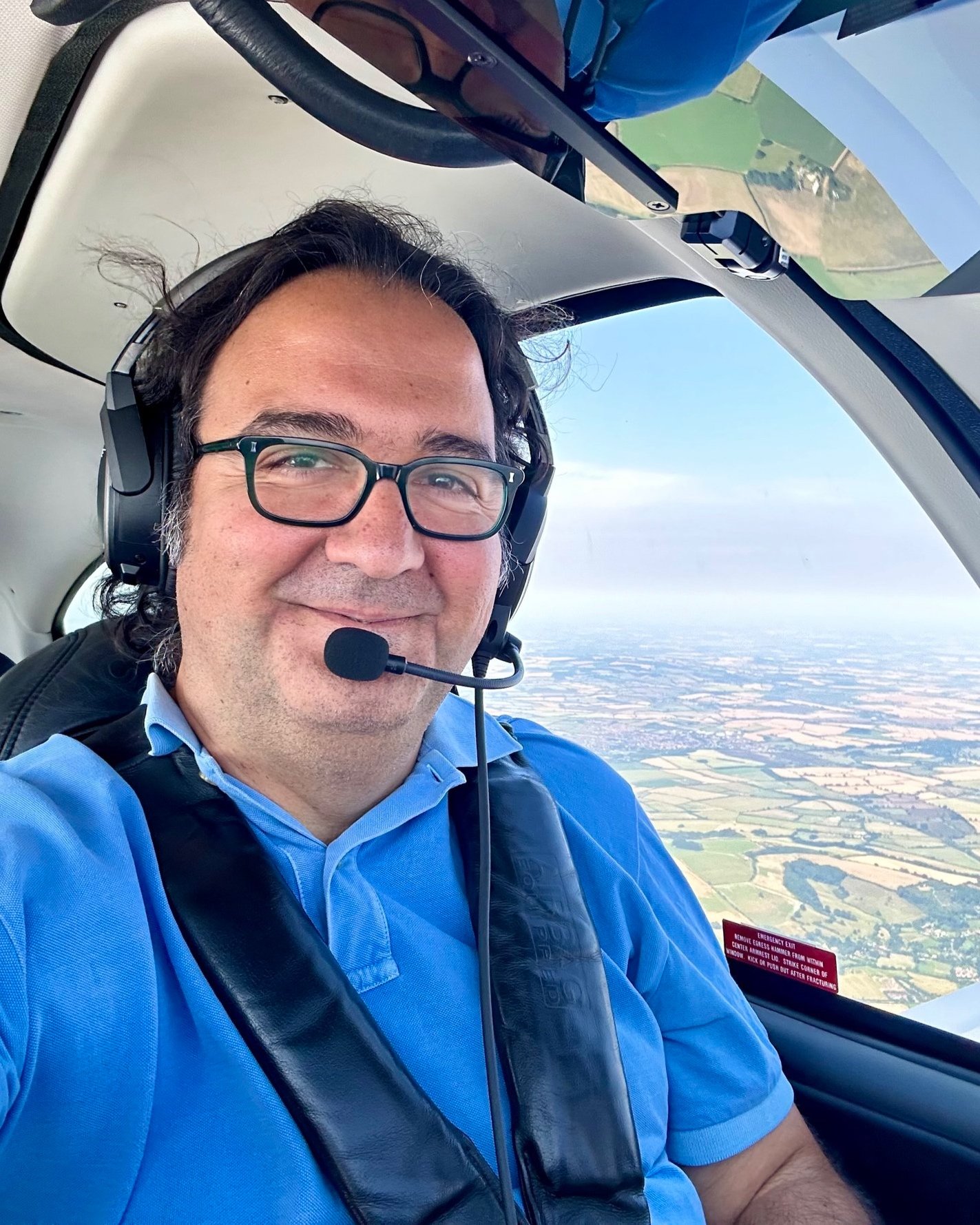 A man with glasses smiling inside a small aircraft cockpit, wearing a blue shirt and headset, with an expansive view of farmland and fields below through the aircraft's window.