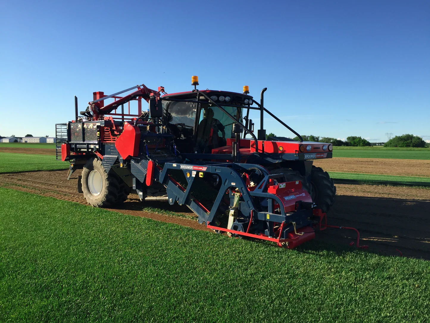 Machine agricole rouge en plein travail sur un champ vert sous un ciel bleu clair.