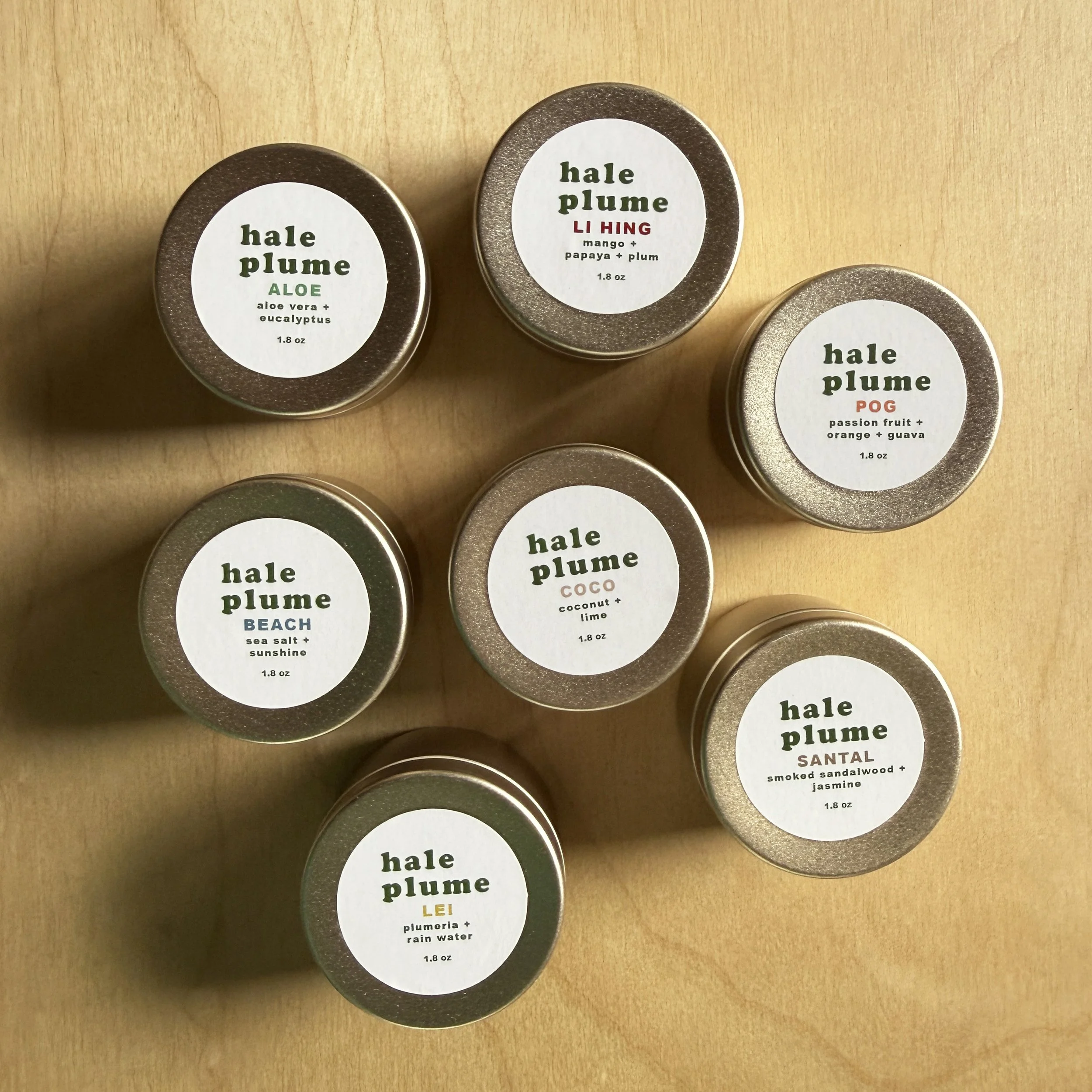 Seven small containers of Hale Plume balm with various scents, arranged in a circle on a white surface with green fern leaves in the background.