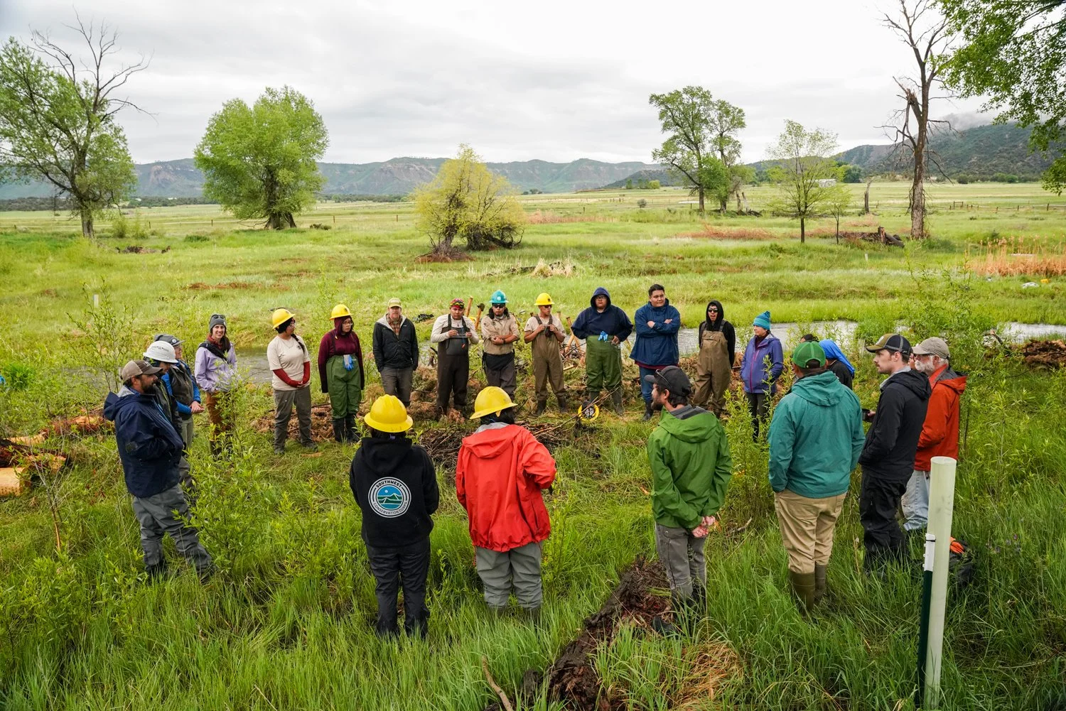 Youth workers and volunteers preparing for a day of woork on the tradtional harvest project in southwest colorado. Everyone is bundled up for the rainy weather and wearing appropriate safety gear. The landscape they are in is very green.
