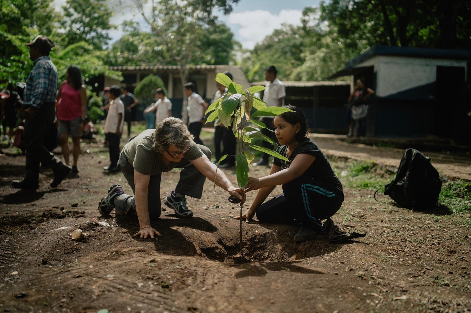 Two women planting a young tree in a hole in the ground while community members gather in the background during a tree-planting event outdoors.