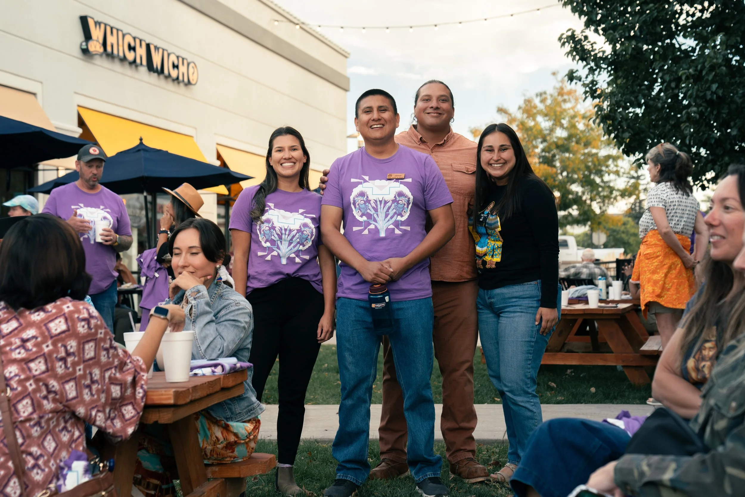 Group of five diverse people smiling and standing together outdoors at a social gathering, with a restaurant called 'High Wicho' in the background and other attendees seated and mingling.