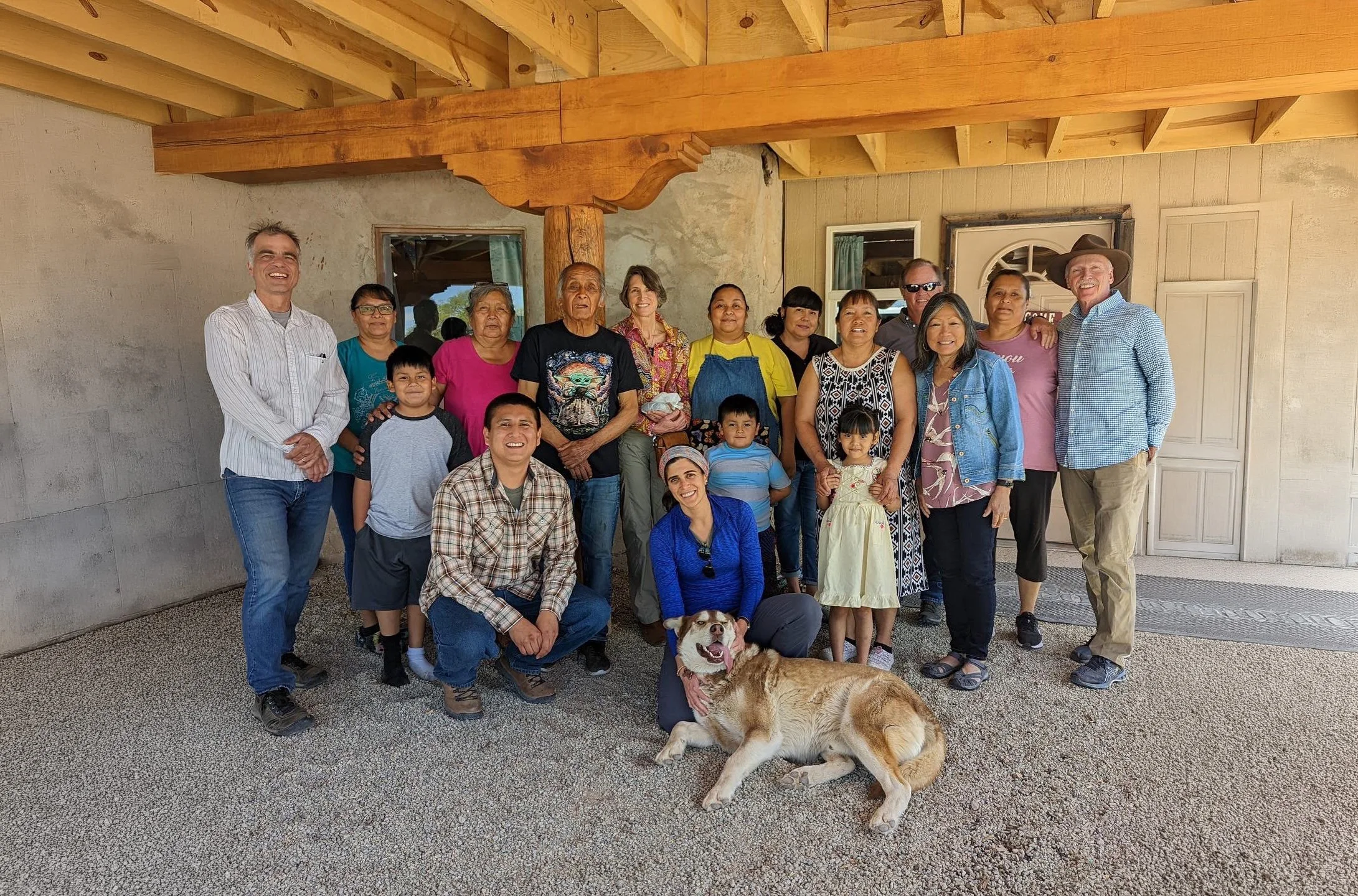 Group of diverse people, including children and adults, smiling and posing together under a wooden structure. There are two dogs, one lying on the ground and one sitting in front of the group. The setting appears to be a porch or patio outside a house.