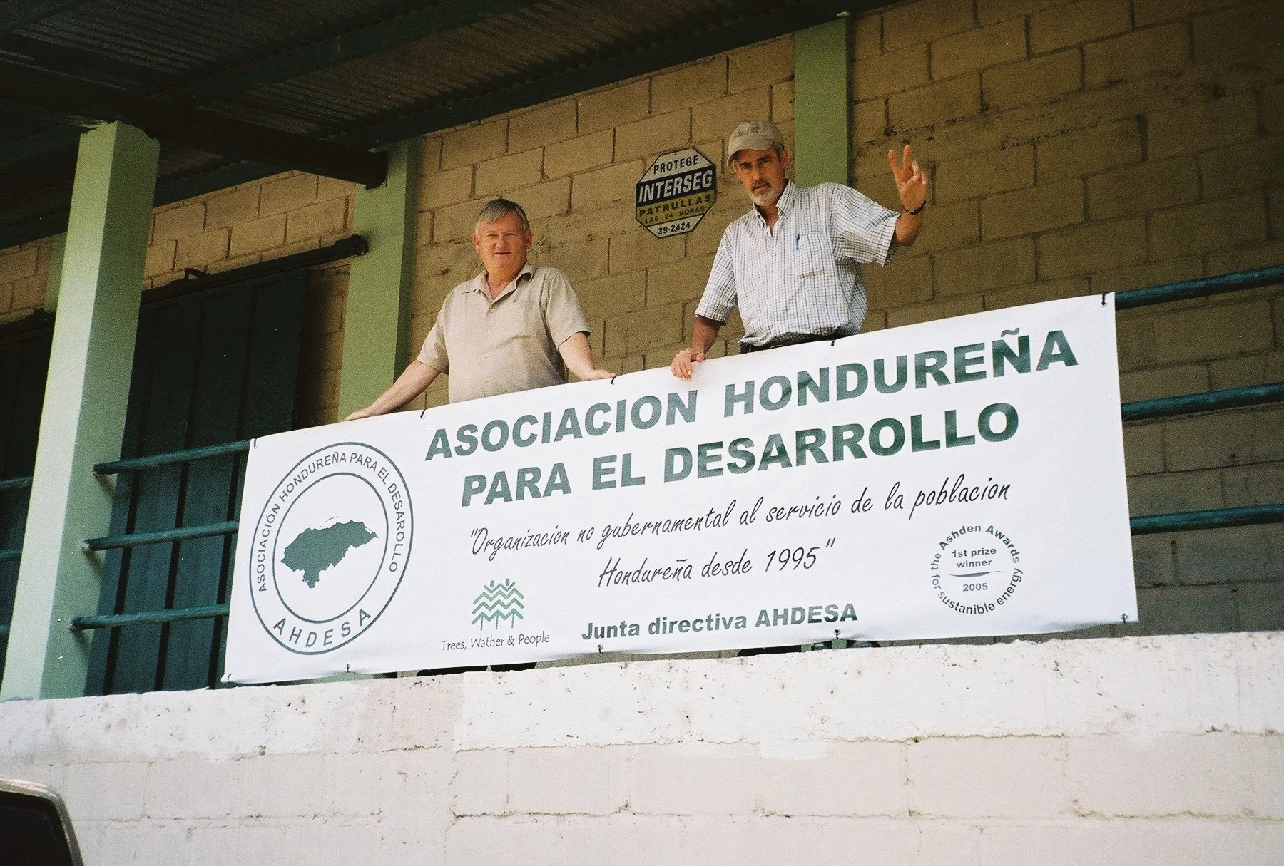 Two men standing behind a banner. The banner reads 'Asociación Hondureña para el Desarrollo' and includes a logo, a quote, and awards. The background shows a brick wall and part of a corrugated metal roof.