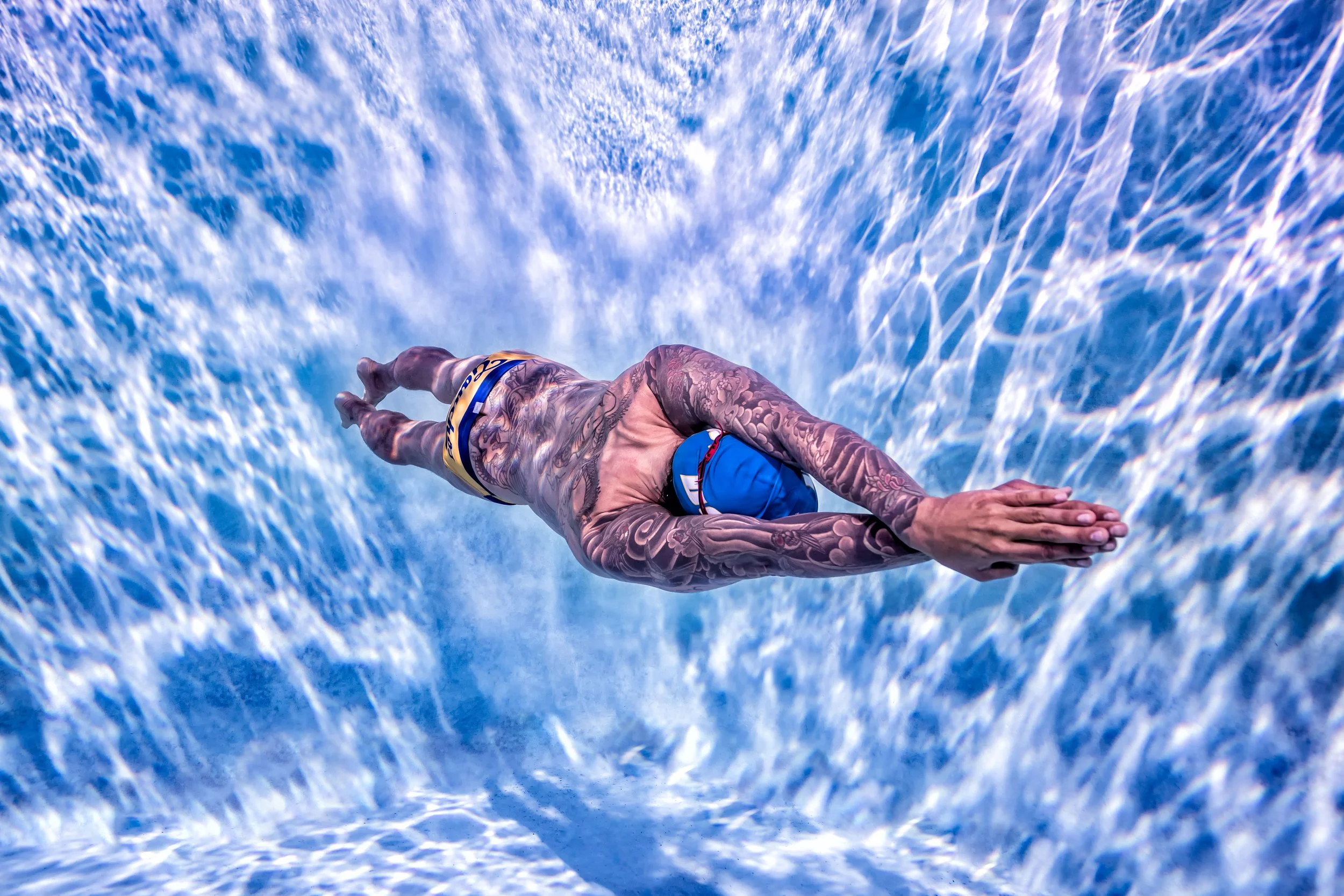 A tattooed person in swimwear appears to be diving into or floating on a water surface with visible light reflections and water movement.