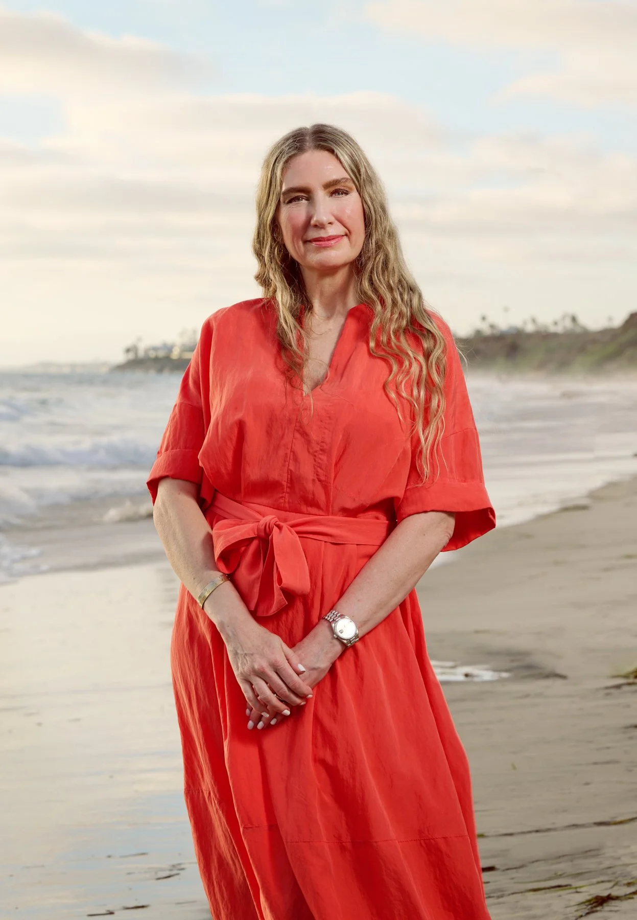 A woman in a red dress standing on the beach with waves and a cloudy sky in the background.