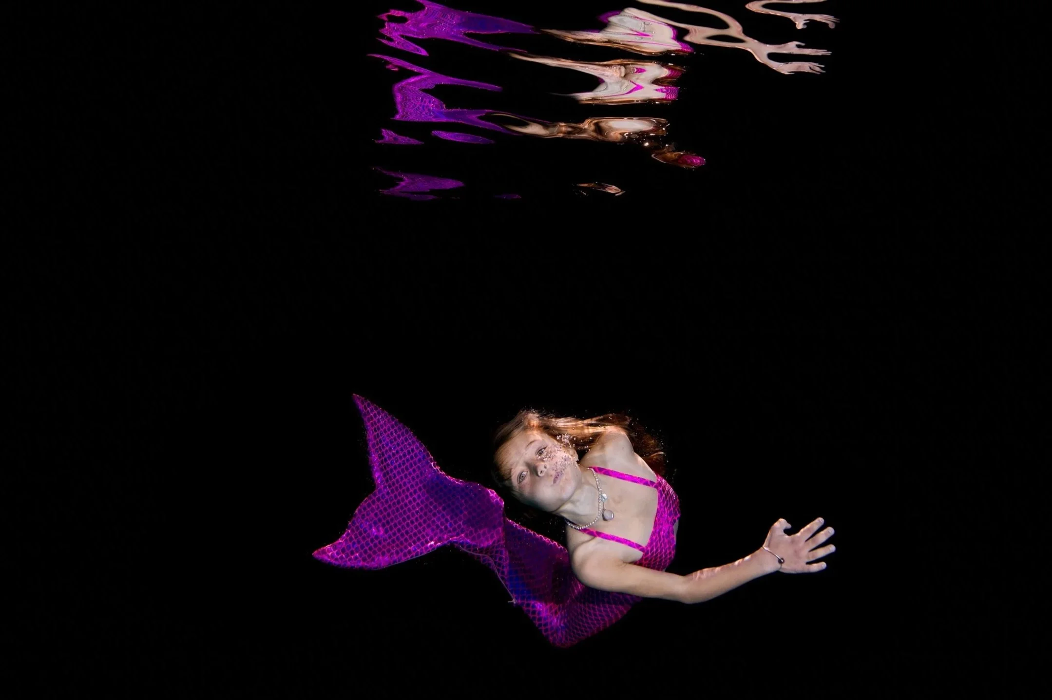 A young girl is lying on her side on a black background, wearing a pink costume with glitter and a purple netted cape, with her head resting on her hand.