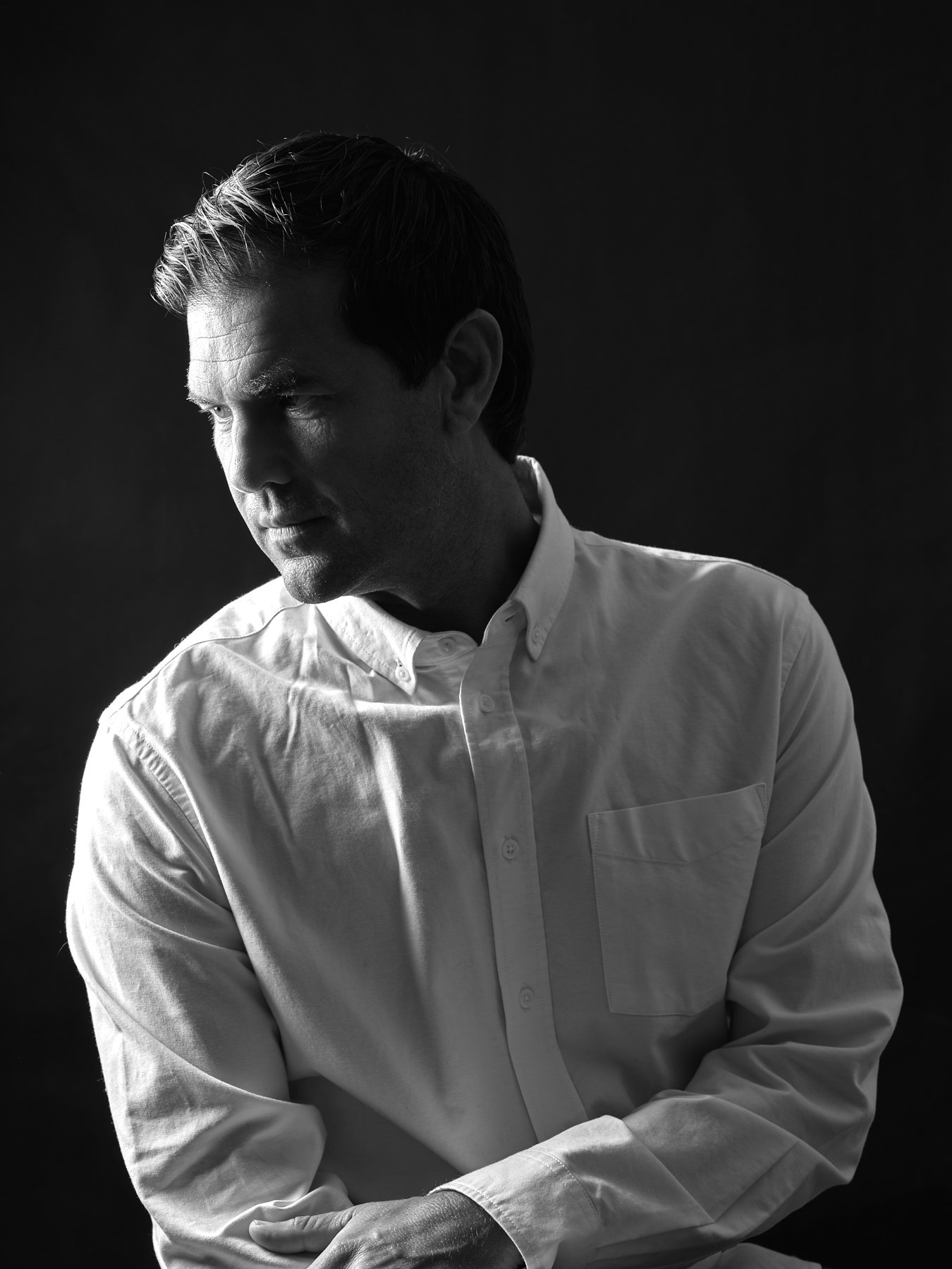A black and white portrait of a man with dark hair in a white button-down shirt, looking downward and to the left, against a dark background.