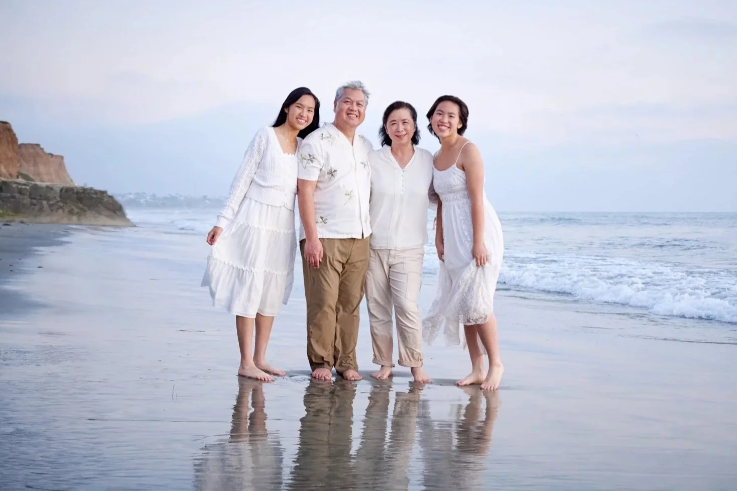 A family of four standing on the beach near the ocean, dressed in white clothing, smiling, with cliffs in the background and waves touching their feet.
