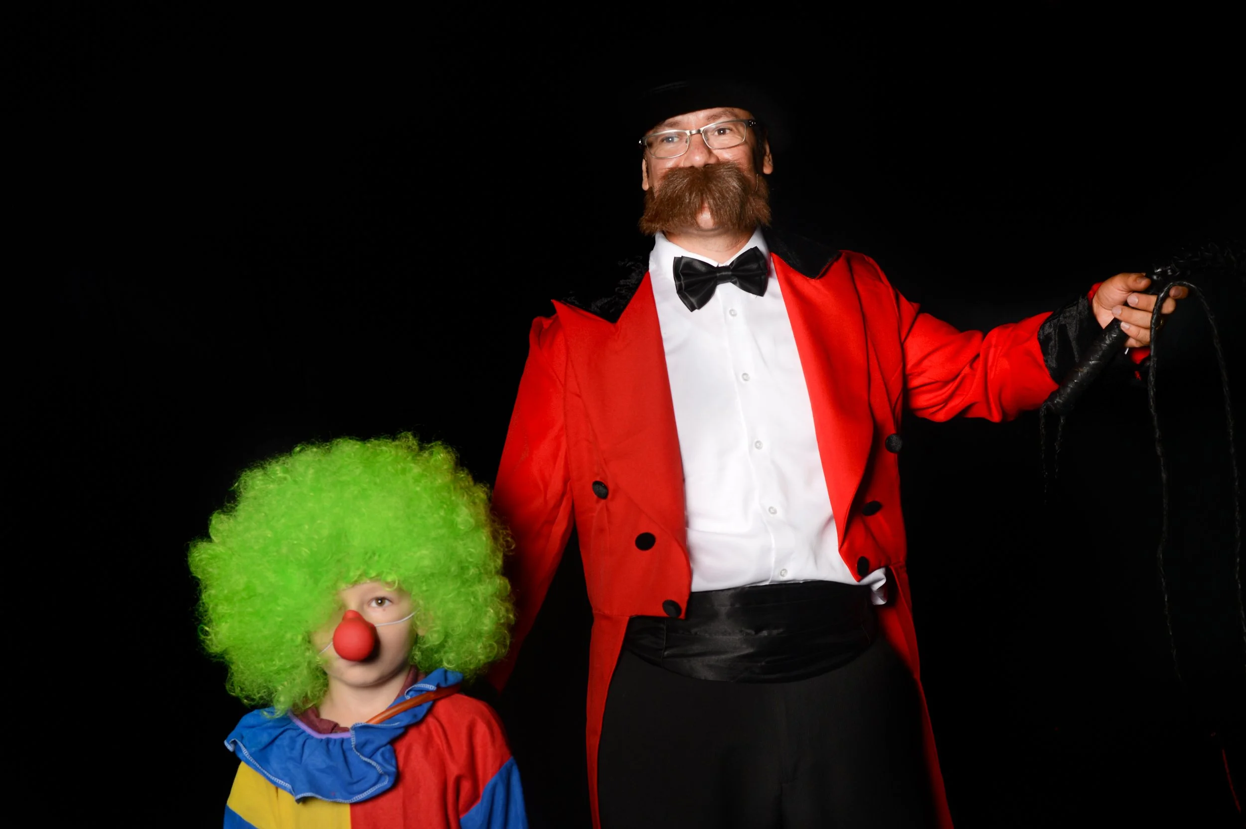 A person dressed as a clown with a large green curly wig, red nose, and colorful costume. Next to them is a man dressed as a magician or ringmaster with glasses, a mustache, a red jacket, black gloves, a white shirt, and a black bow tie, holding a wh