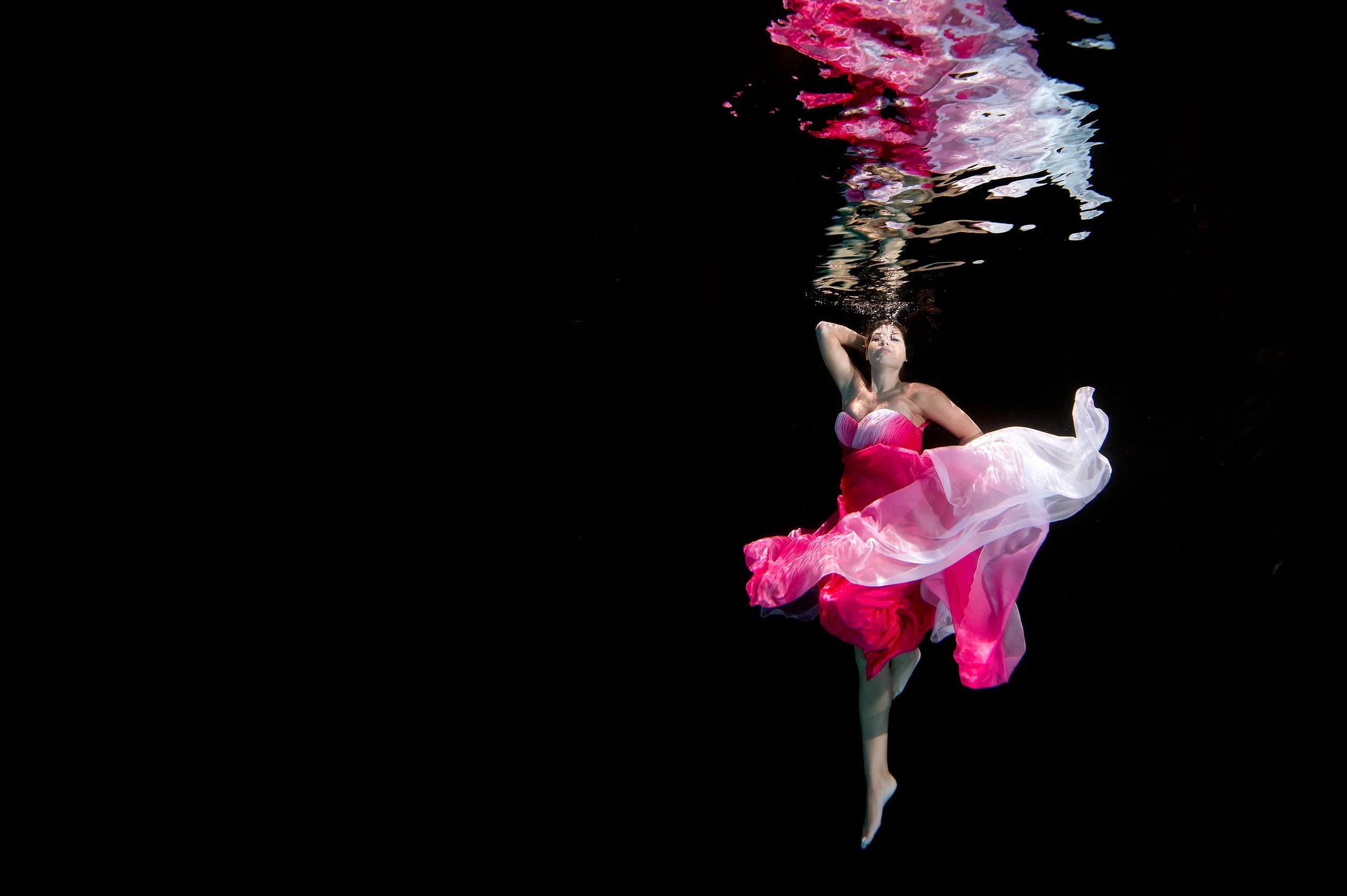 A woman in a pink dress is floating in dark water, with her hand near her face and her eyes closed. Her reflection is visible on the water's surface.