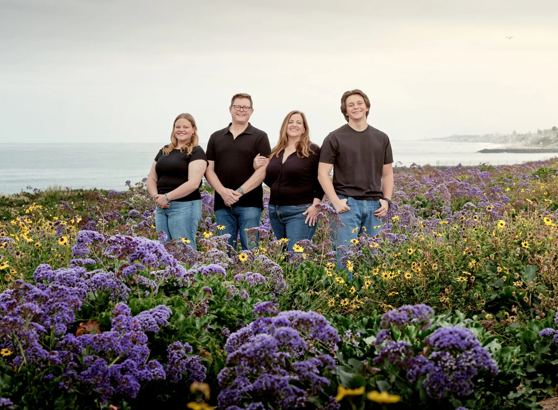 Four people standing on a field of purple and yellow flowers near the coast, smiling and posing for a photo.