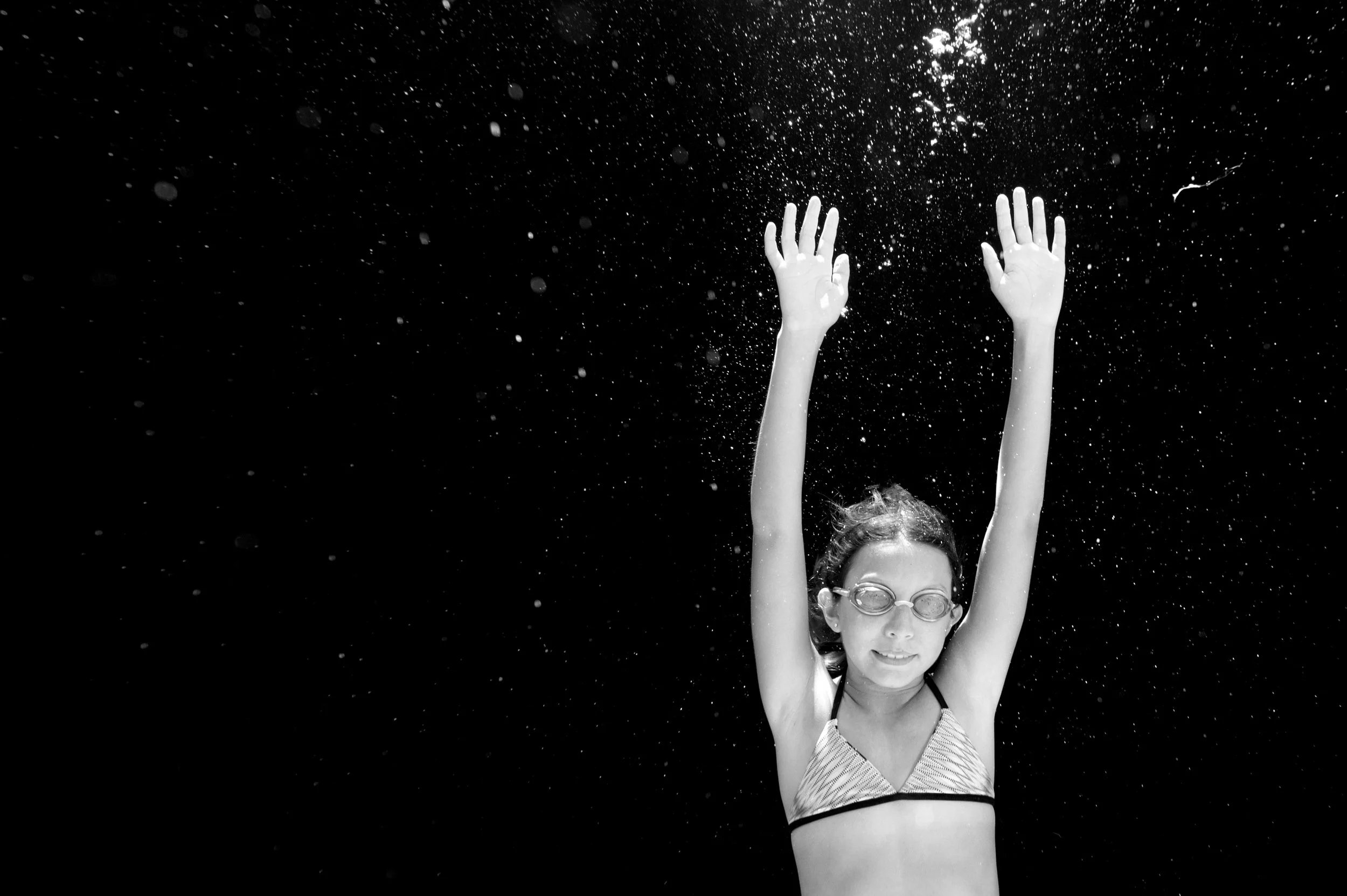 A woman wearing glasses and a striped bikini top is submerged underwater with her arms raised above her head, surrounded by water bubbles.
