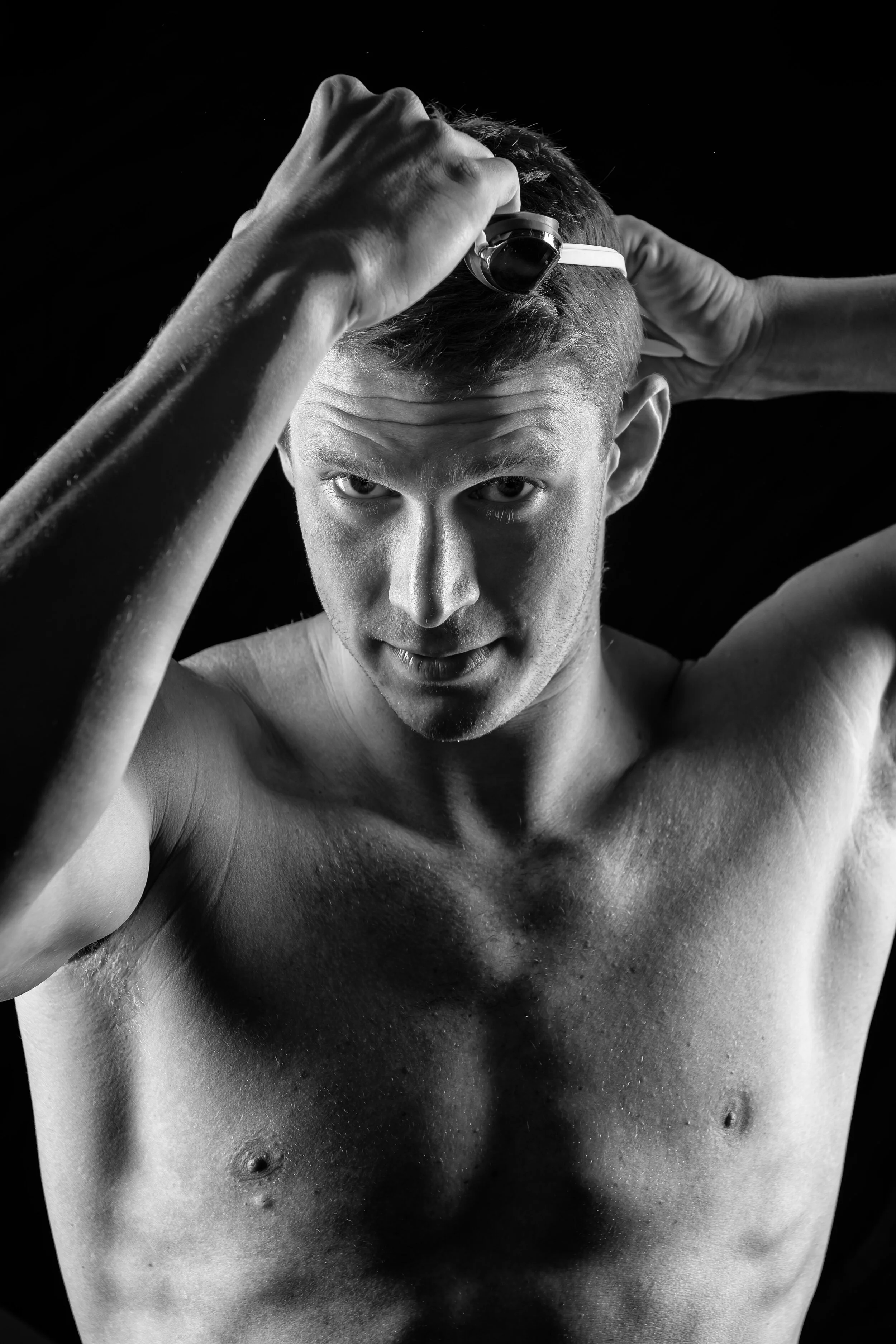 Black and white photo of a shirtless young man with short hair, looking into the camera, with his hands on his head, against a dark background.