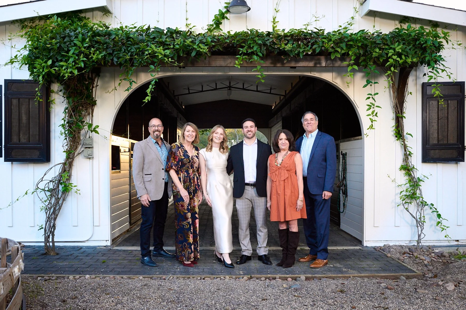 Group of six adults standing in front of a white building with an arched entryway and greenery, dressed in formal and semi-formal attire, smiling for the photo.
