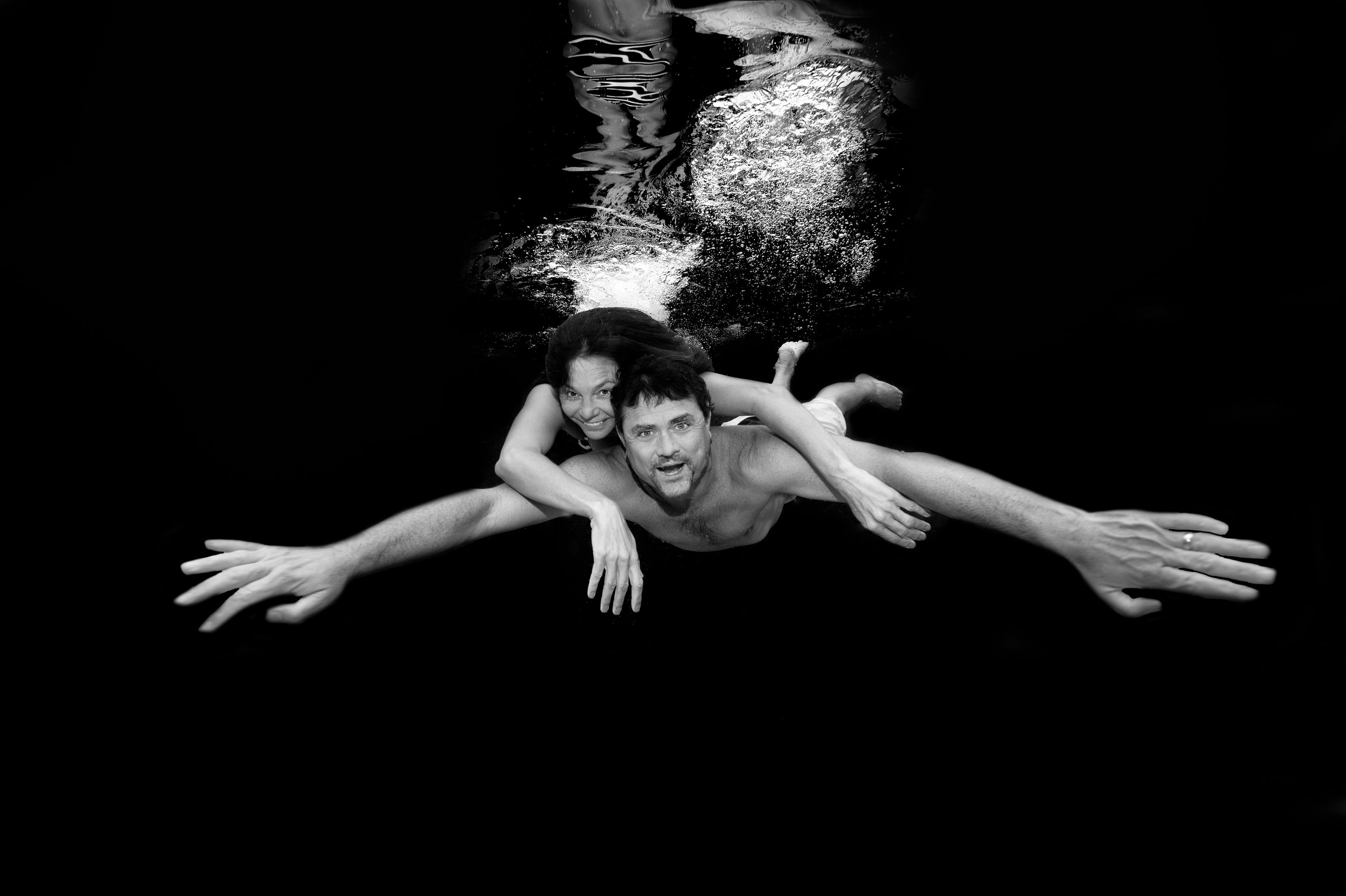 Black and white photo of a man and woman swimming underwater, facing the camera, with the woman's arms around the man's neck, both smiling.