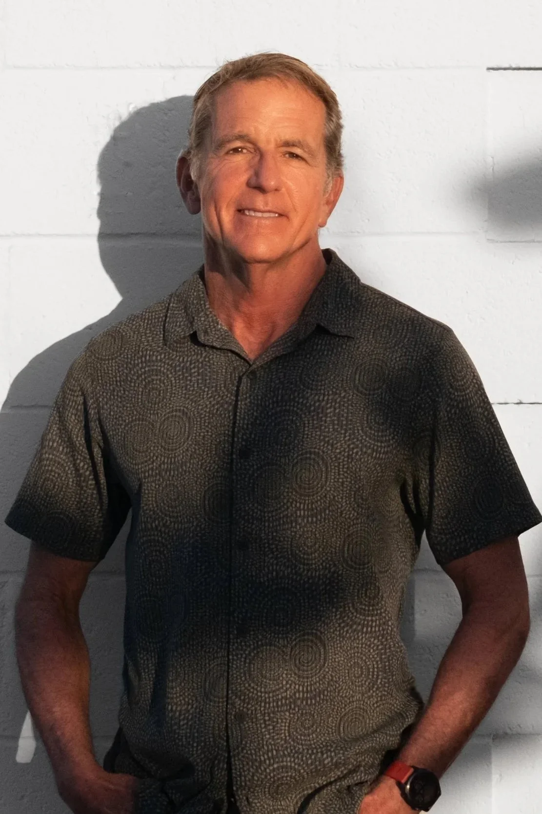 A middle-aged man in a patterned short-sleeve shirt standing against a white wall with a shadow cast behind him.