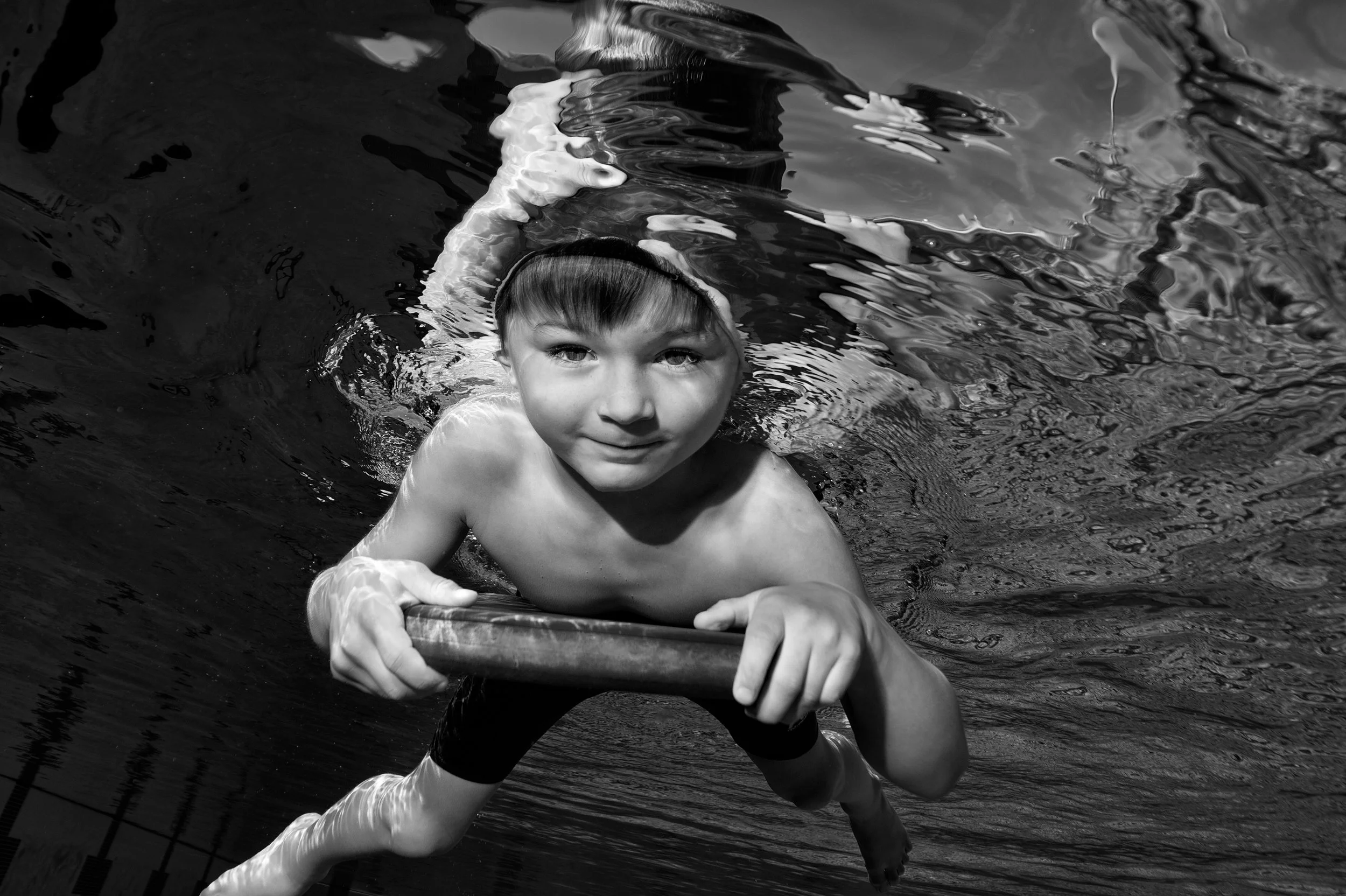 A young boy swimming in a pool, holding onto a pool float with both hands, with water ripples around him.